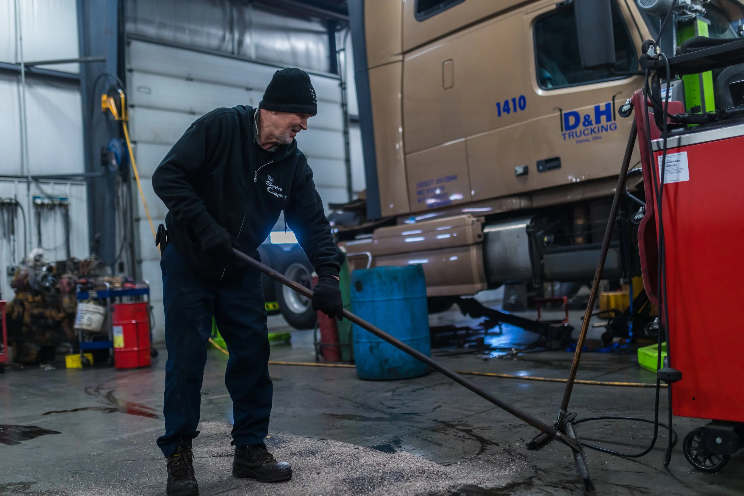 A worker in a black jacket and beanie cleans the concrete floor in a garage or workshop with a power scrubber. A large semi-truck is visible in the background with the logo 'D&H TRUCKING' and the number 1410.