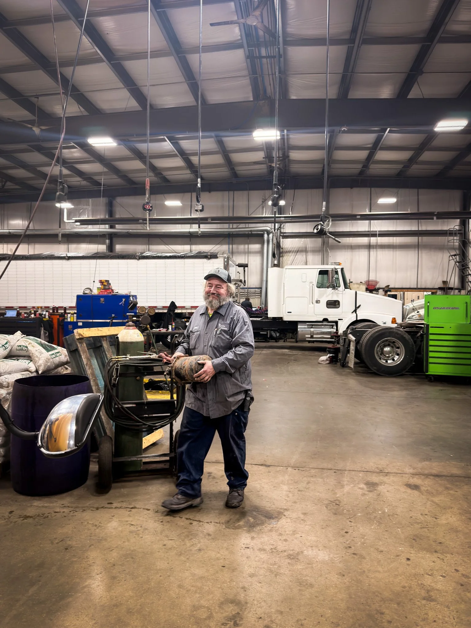 A smiling man with a beard, wearing a gray uniform and cap, standing in a spacious industrial workshop. He is holding what appears to be a heavy metal object, surrounded by various tools and equipment, with trucks and machines in the background.