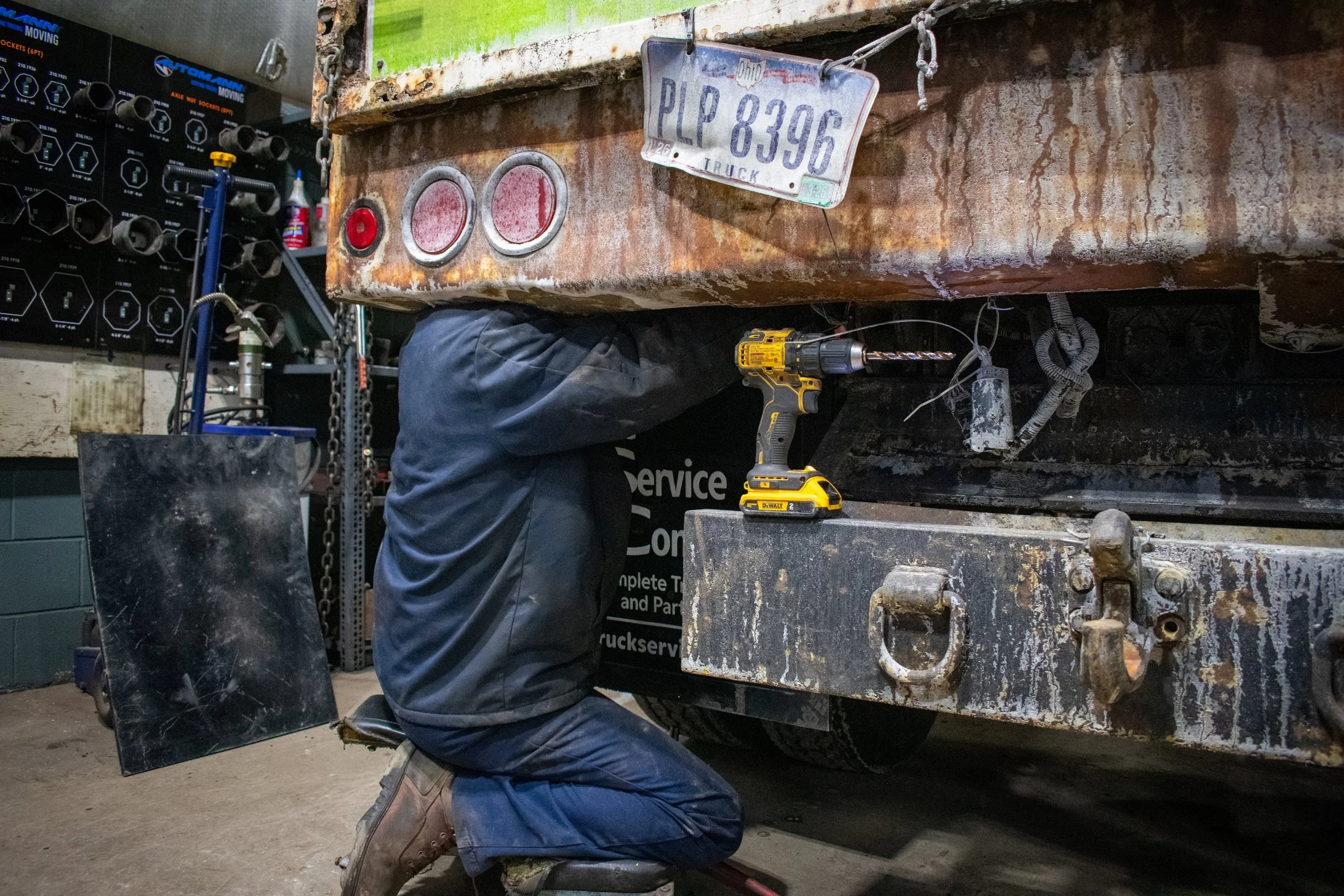 A mechanic kneeling and working underneath a rusty, green truck with a yellow drill on the ground nearby.