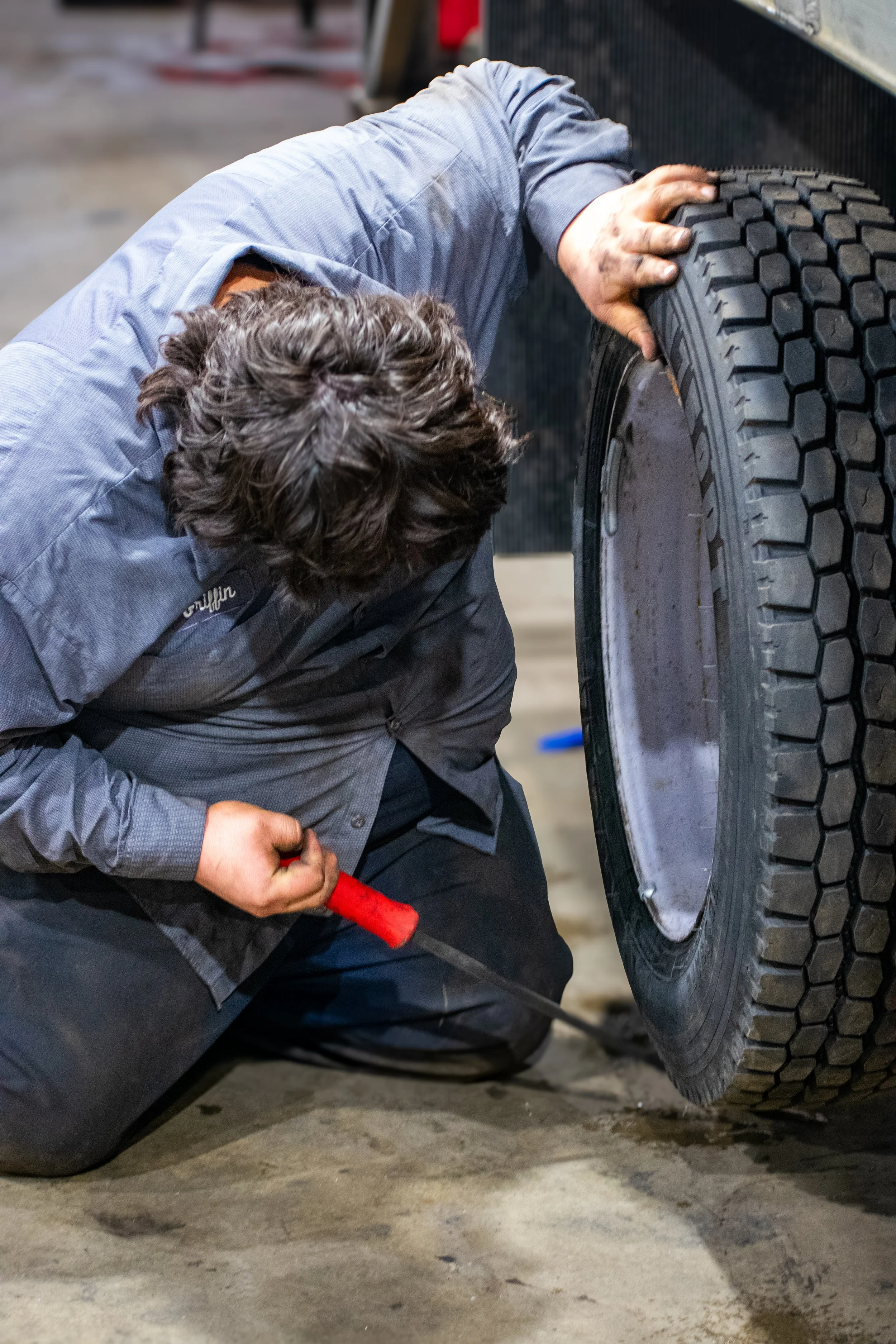 A person kneeling on the ground, repairing or inspecting a large tire of a vehicle, using a screwdriver in a workshop or garage setting.