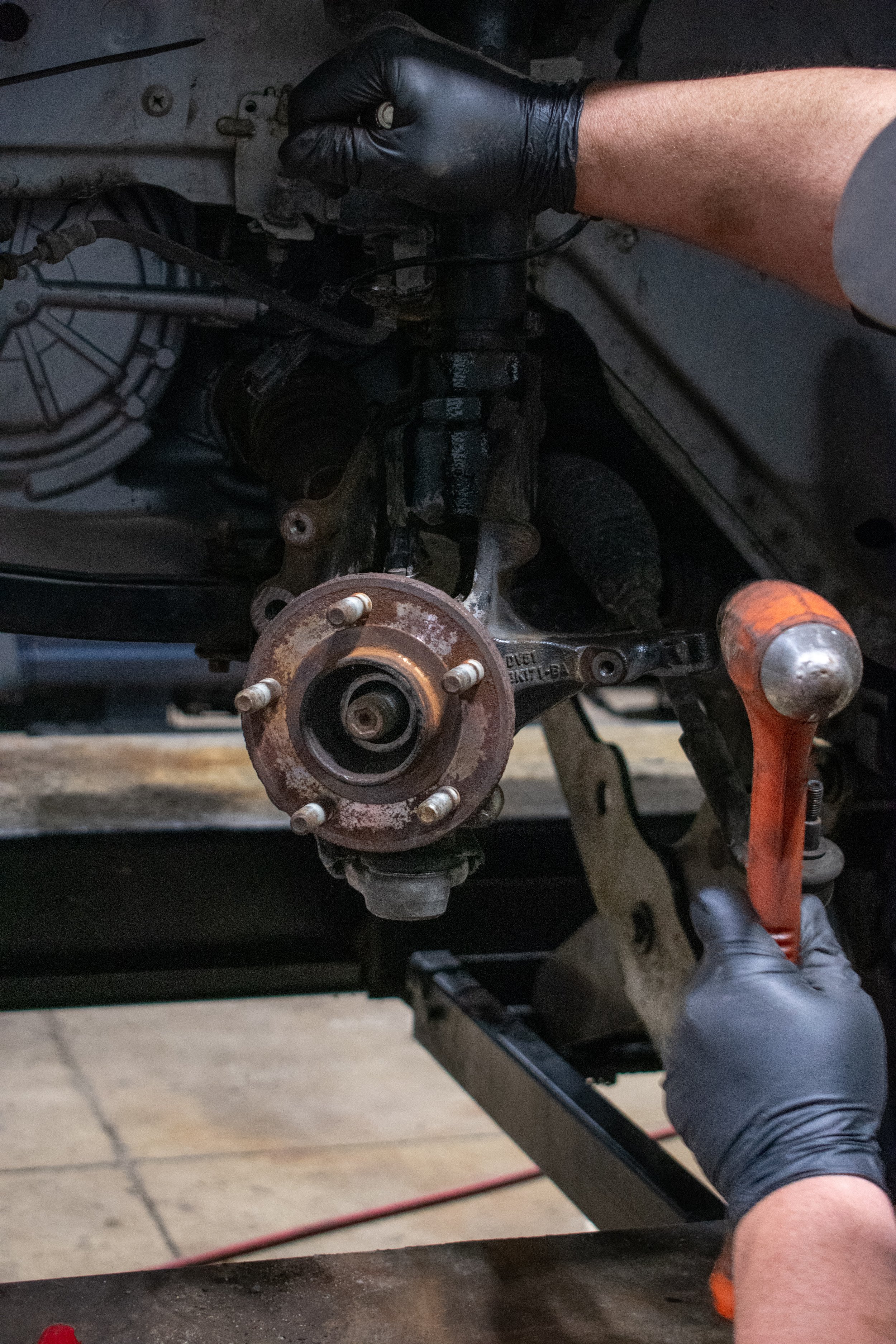 A mechanic wearing black gloves using a crowbar to work on a disassembled car suspension system, with the hub and wheel removed.
