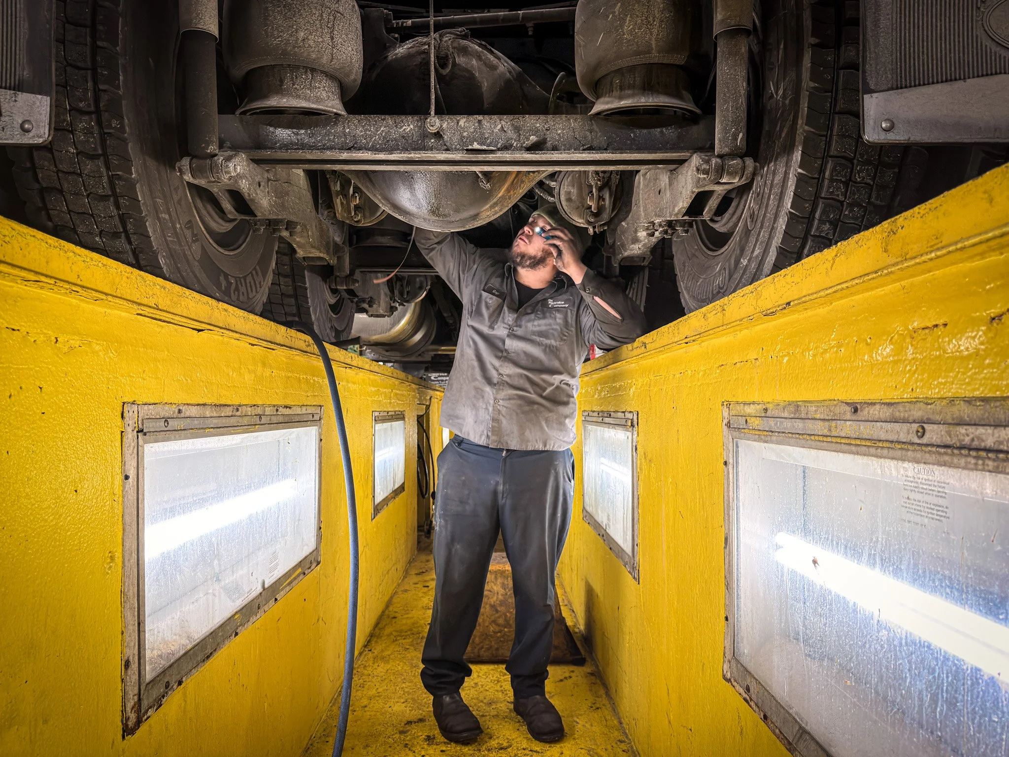 A mechanic inspecting the underside of a vehicle on a yellow inspection lift. The mechanic is using a tool and wearing a gray jumpsuit.