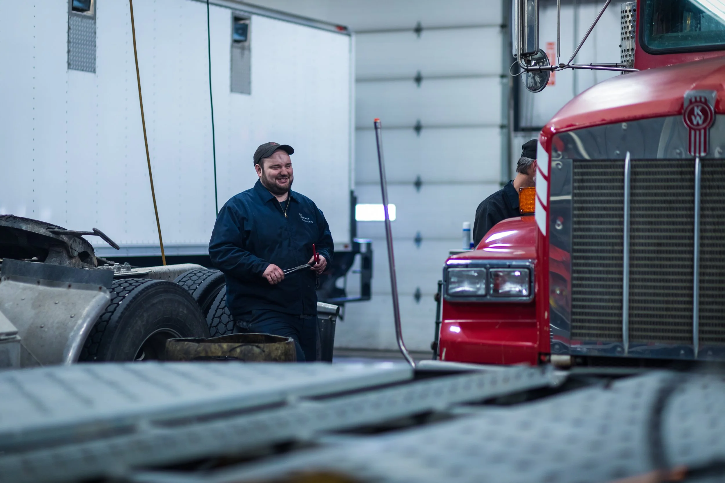 Two mechanics working inside a vehicle service garage, one smiling and holding a tool, with a red semi-truck and a white trailer in the background.