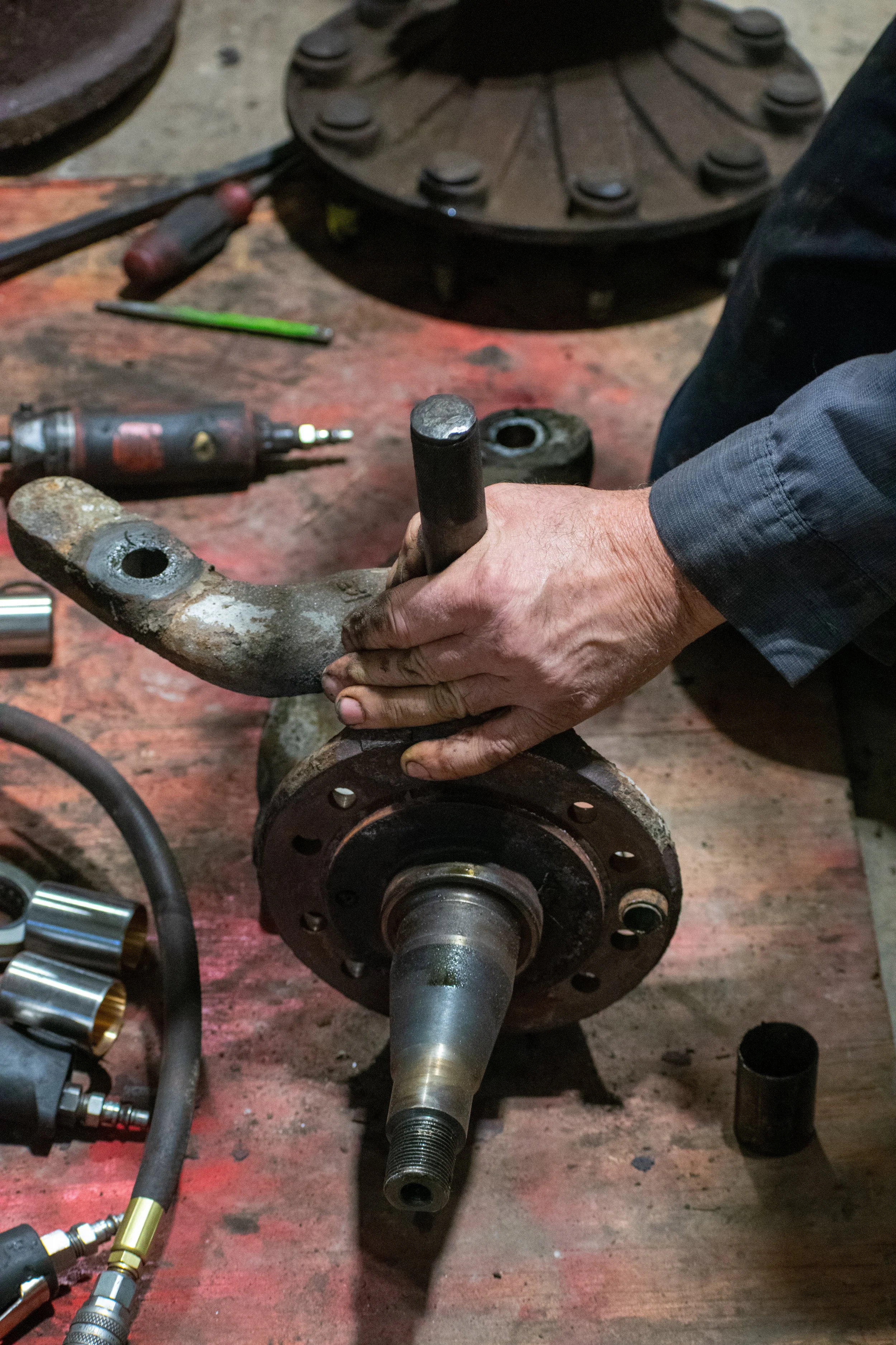 A person working on a rusty automotive part, likely a wheel hub or spindle, on a workbench with tools and parts around.