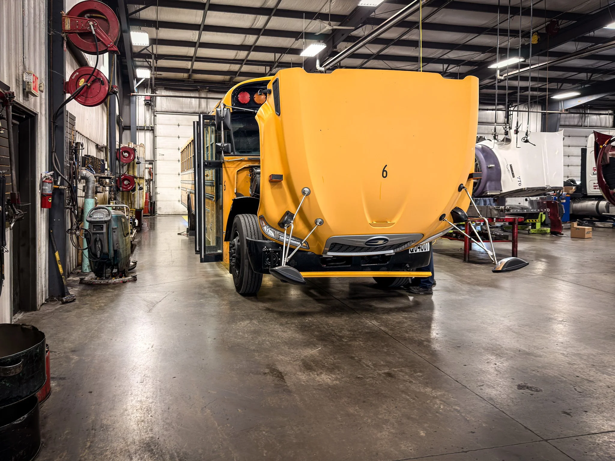 Inside a workshop containing a yellow bus with its front facing the camera, positioned in the center and partially lifted. The workshop has various tools and equipment on the walls and floor, including fire hoses, a machine, and large industrial lights.