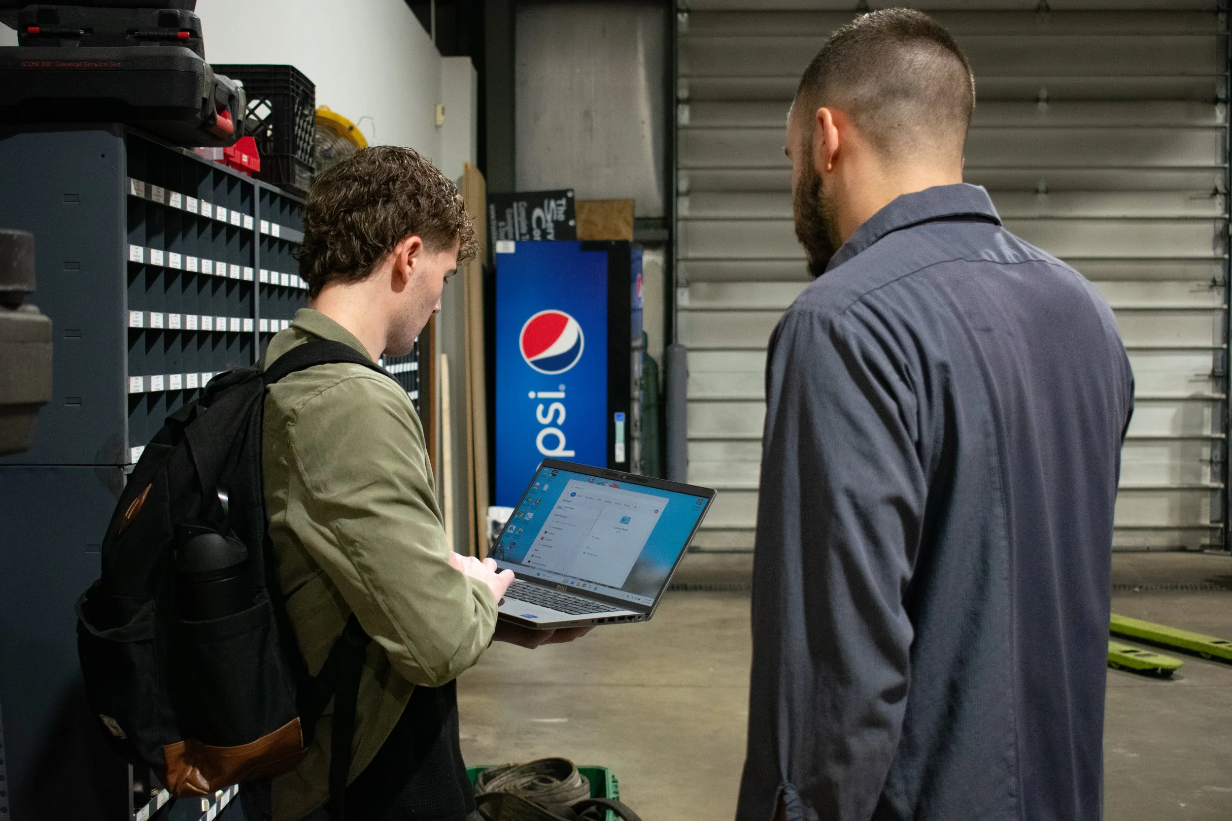 Two men in a warehouse or garage, one with a backpack, looking at a laptop screen while the other stands beside him. There is a Pepsi vending machine and metal garage door in the background.
