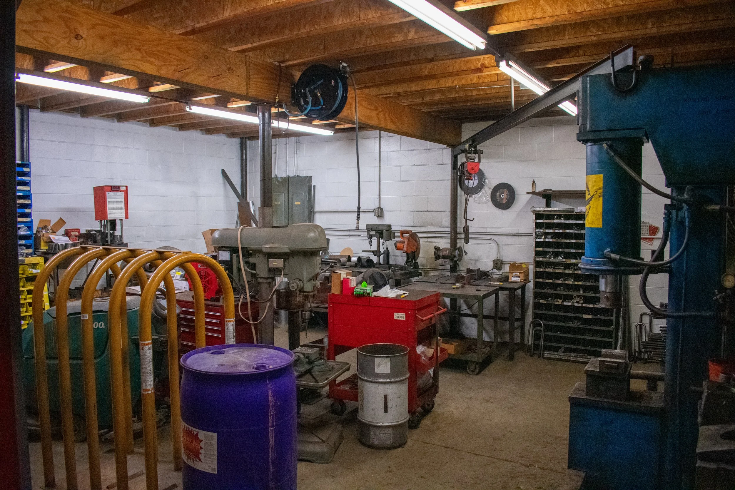 Industrial workshop with various tools, equipment, and storage units, including a drill press, metal shelves, and a red tool chest, illuminated by fluorescent lights.