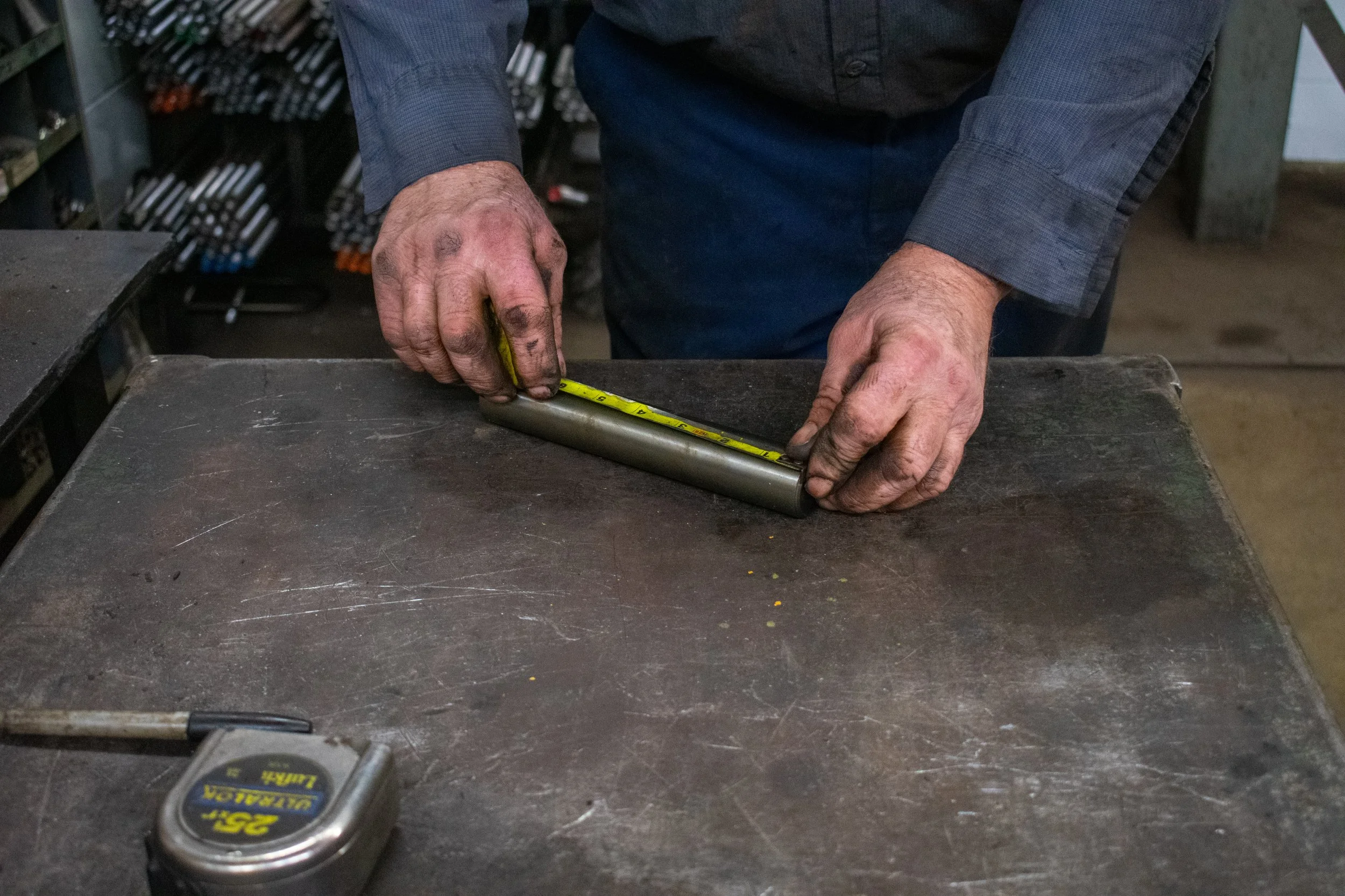 A person measuring a metal pipe with a yellow tape measure on a workbench in a workshop.