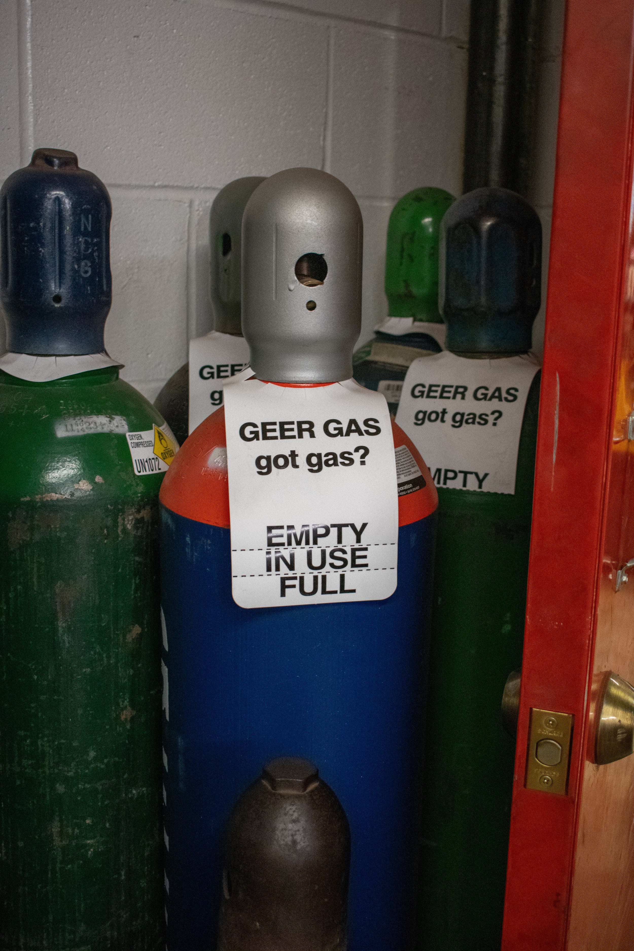 Gas cylinders stored in a utility closet, with a sign on one indicating they are empty but in use, and prompting 'GEEK GAS got gas?'