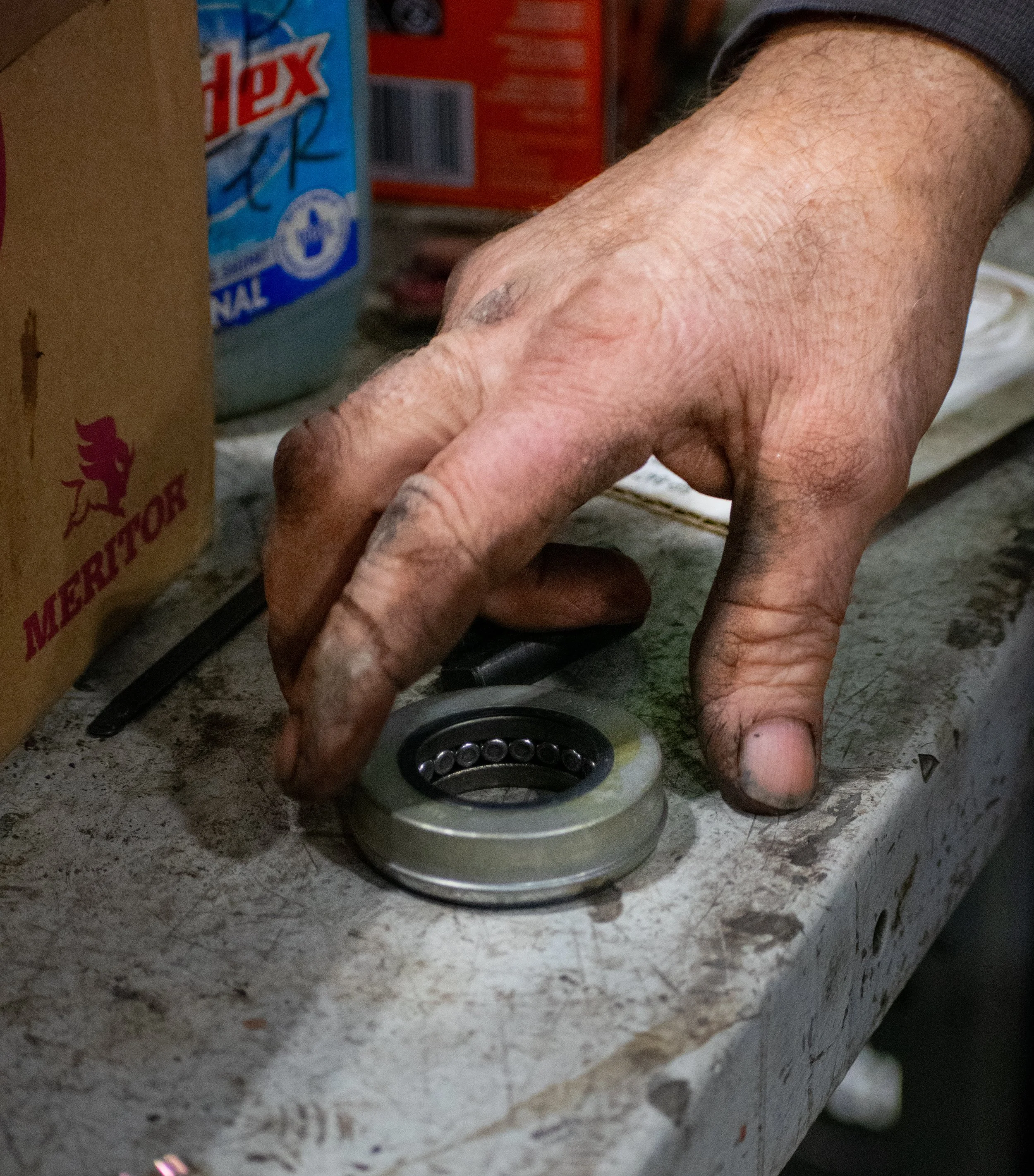 A hand, covered in dirt and grease, holds a metal ball bearing on a dirty workbench with various tools and boxes in the background.
