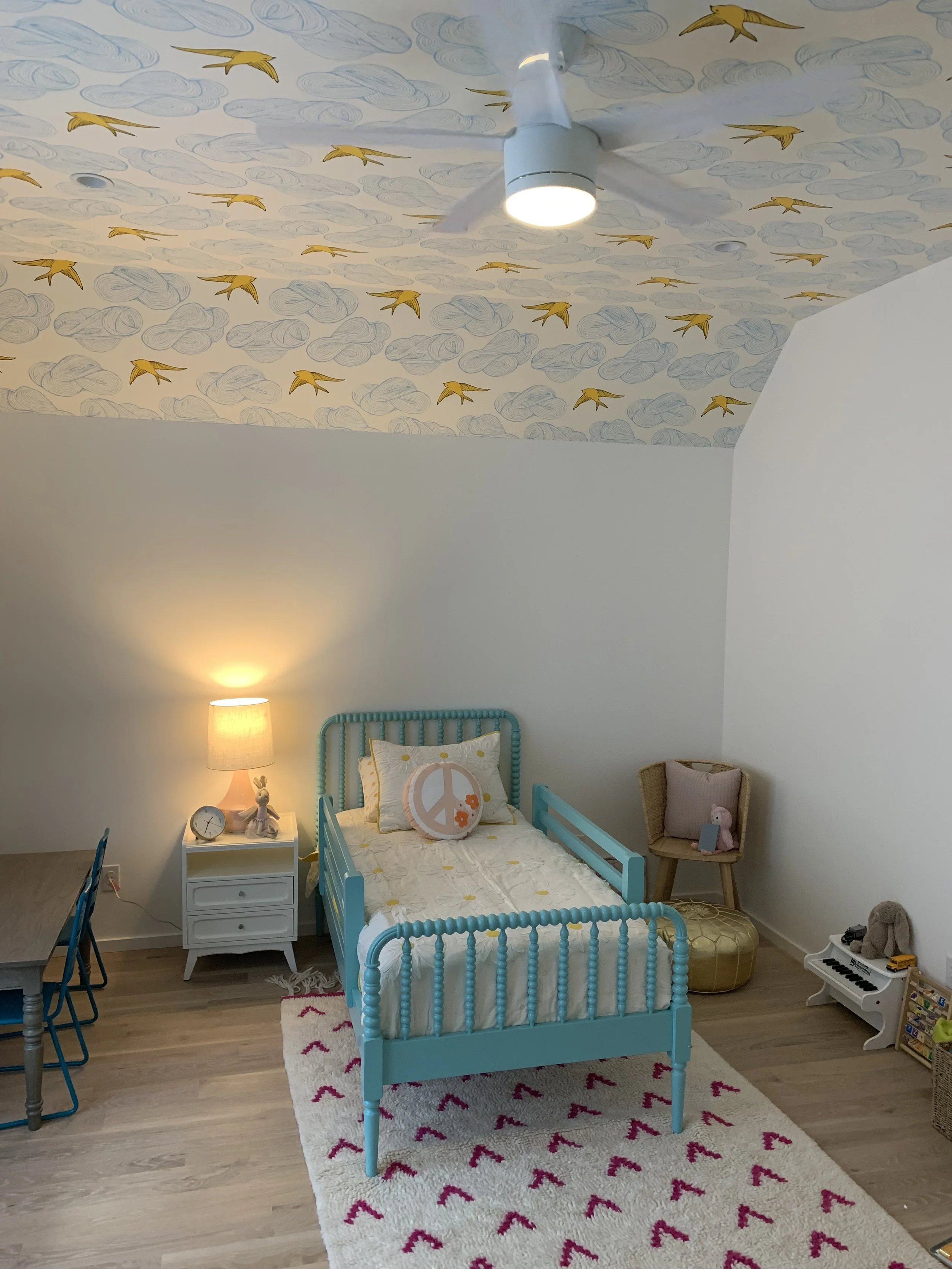 Child's bedroom with a turquoise bed, a white nightstand with a lamp, a pillow with a peace sign, a stuffed animal, a small piano, and a decorative wall with clouds and yellow birds painted on the ceiling.