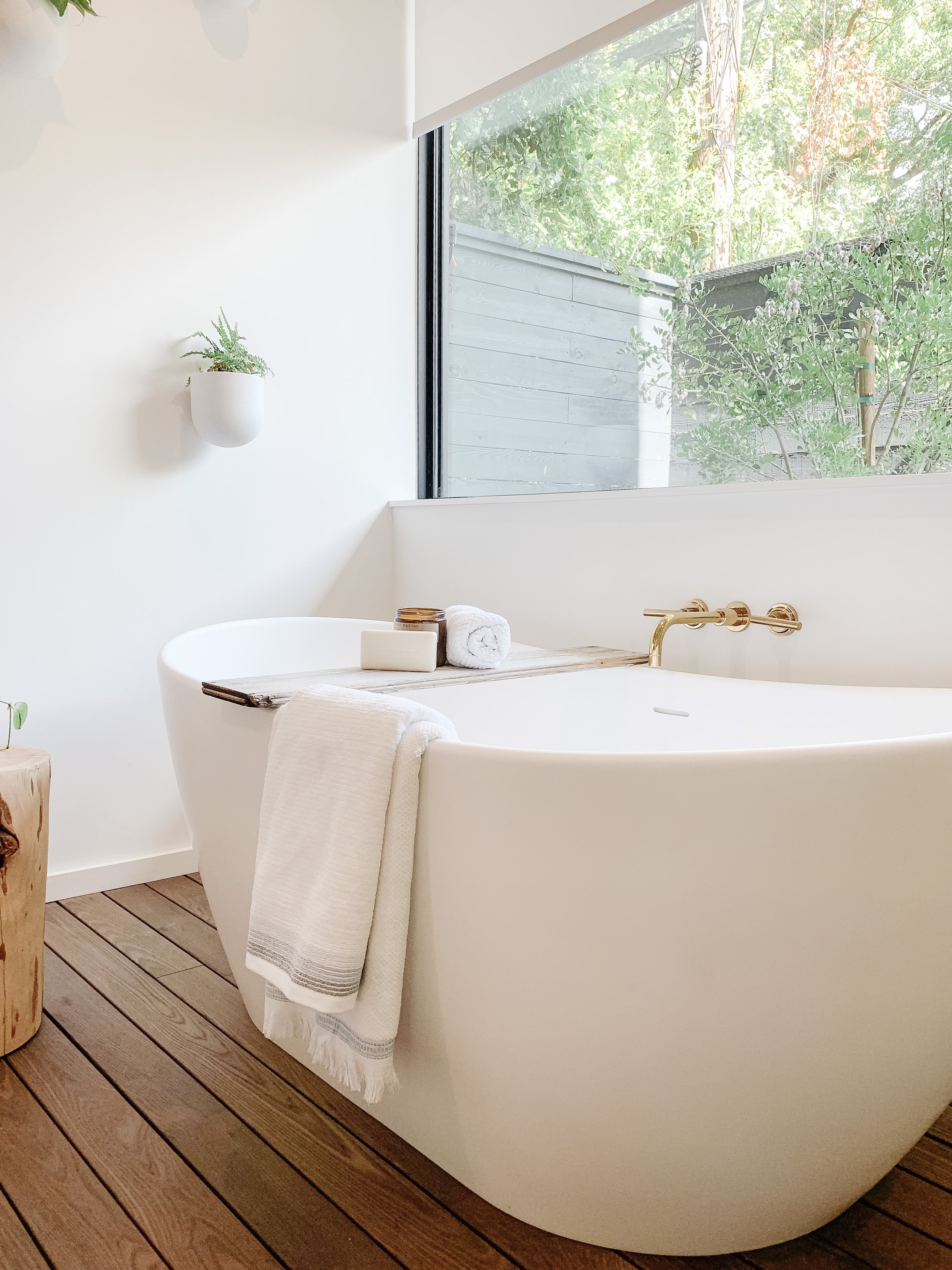 Modern bathroom with white freestanding bathtub near a large window, wooden floor, and minimalistic decor including a wall-mounted plant and bath essentials.