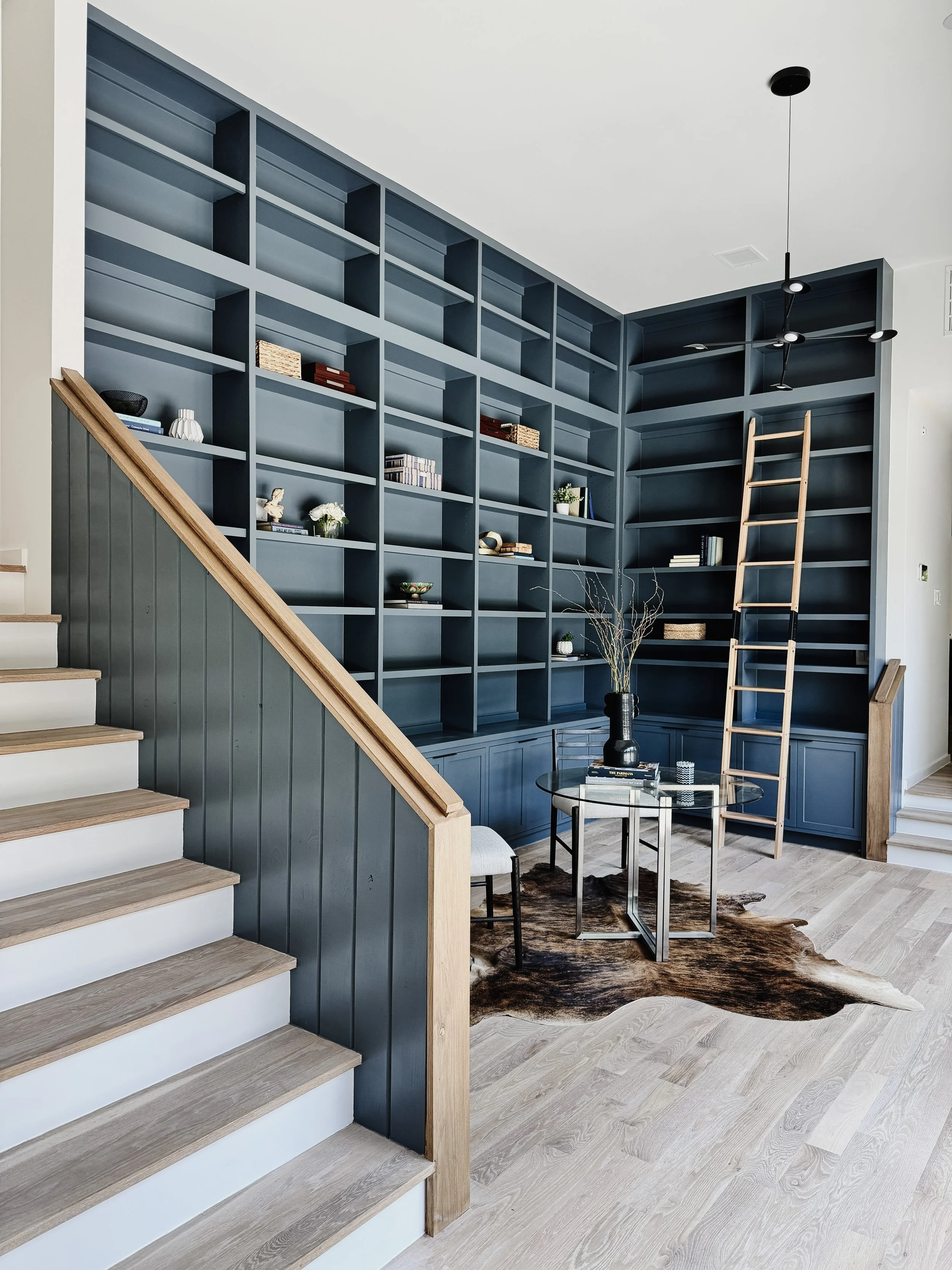 Interior view of a room with a large blue bookshelf, a ladder, a small sitting area with a glass table, a chair, and a cowhide rug.