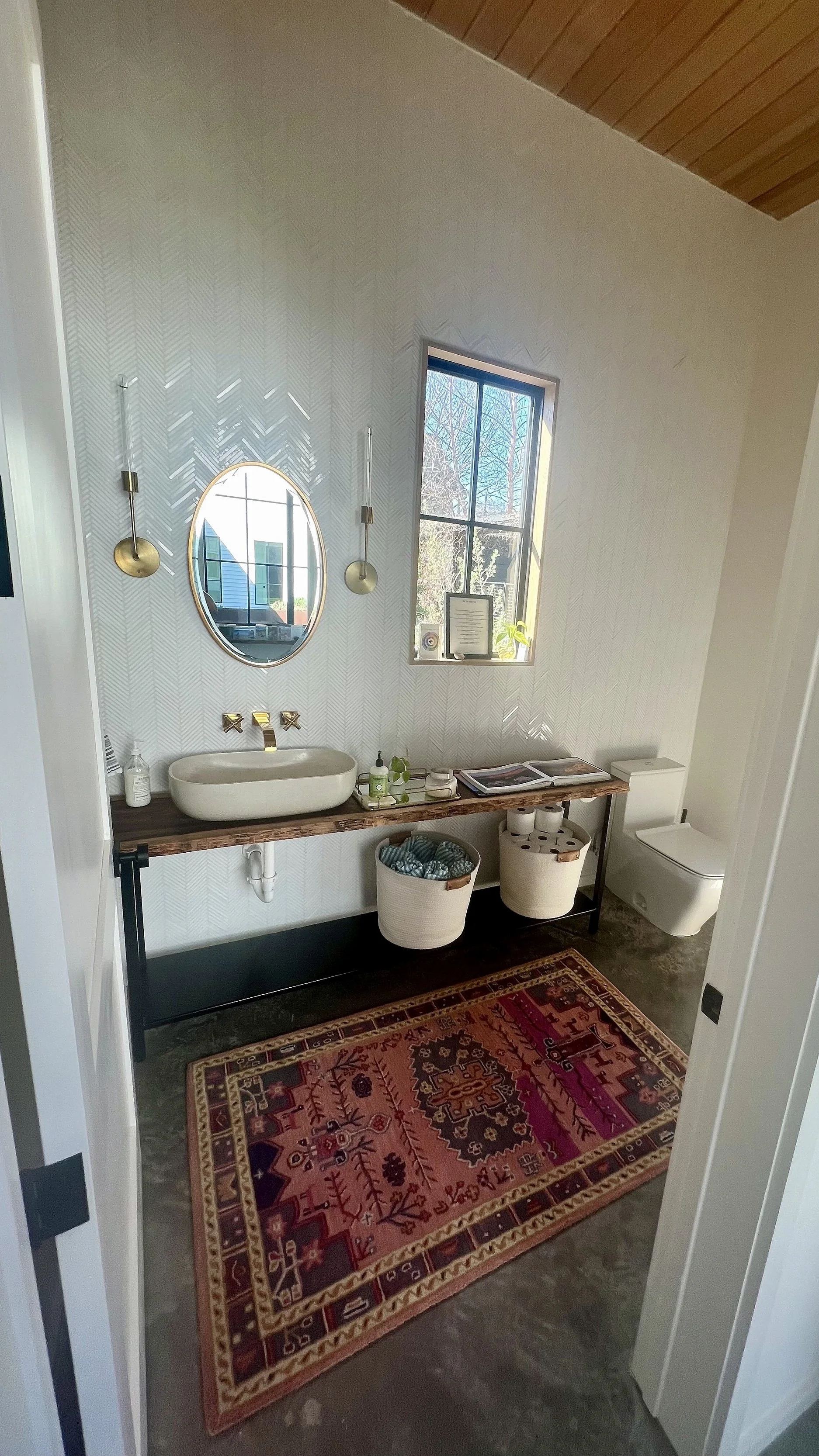 A modern bathroom with a wooden vanity, oval vessel sink, and brass fixtures. A coral-colored patterned rug lies on a concrete floor, and there's a window with a small plant and framed certificate on the sill.
