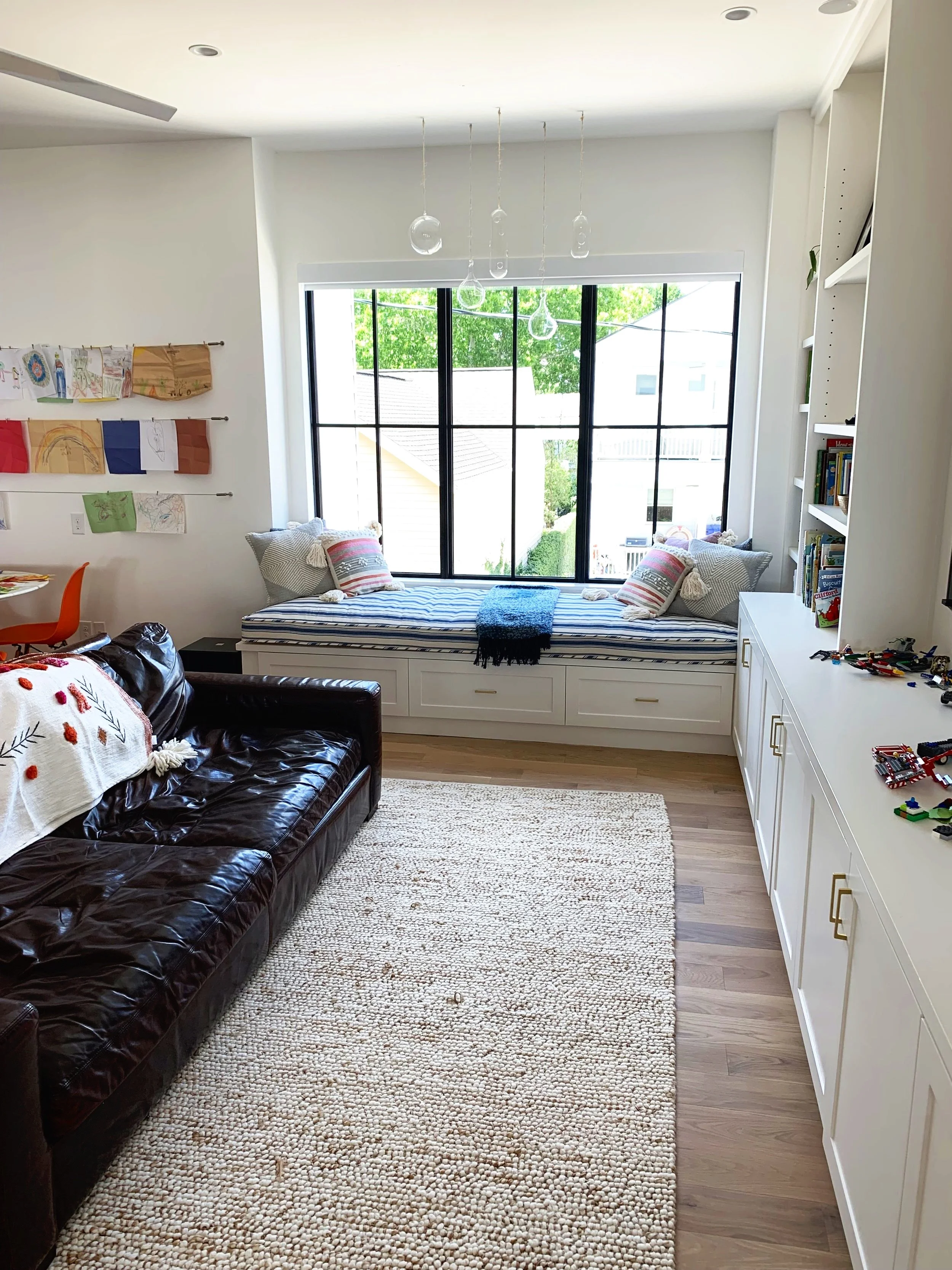 A room with a large window seat under a black-framed window, a dark brown leather sofa with a white blanket, a white rug, and a white cabinetry with books and toys, decorated with hanging glass light fixtures and artwork on the wall.