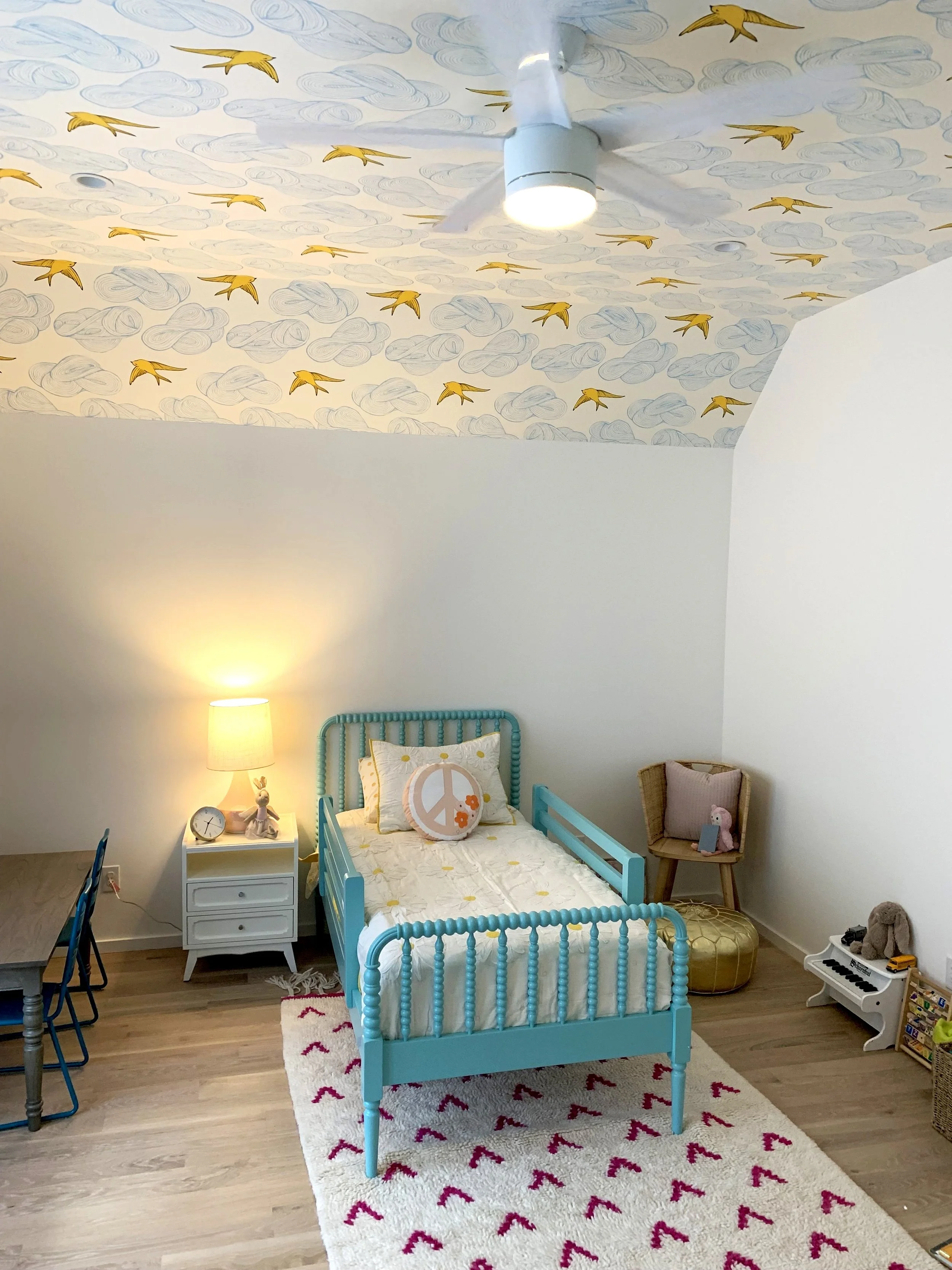 Children's bedroom with a blue bed, peace sign pillow, stuffed animals, a small white nightstand with a lamp and clock, a colorful rug, and a ceiling decorated with birds and clouds mural.