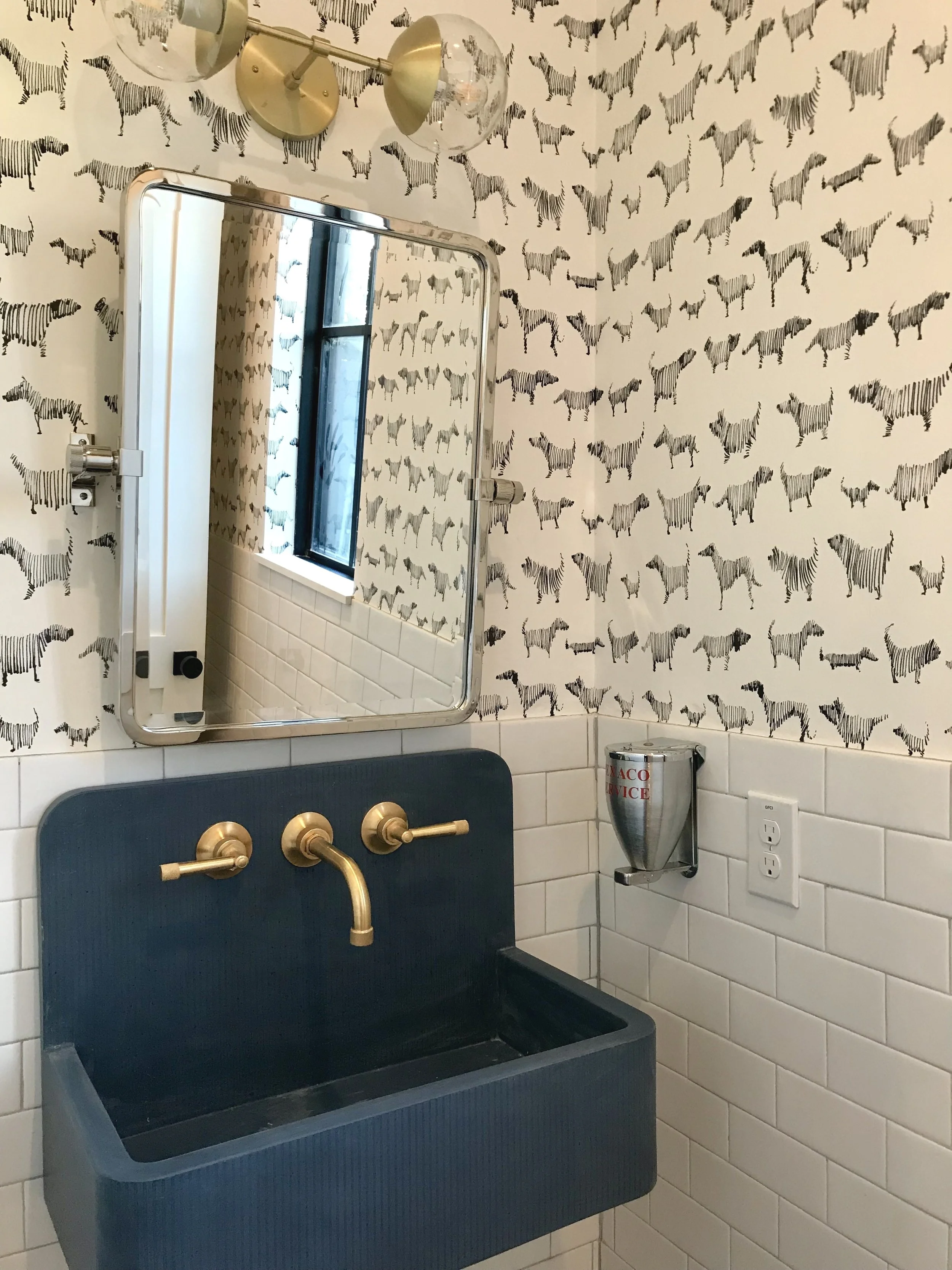 A bathroom with black and white zebra patterned wallpaper, a gold-lit wall-mounted light fixture, a rectangular mirror, a black sink with gold fixtures, a wall-mounted soap dispenser, and a window with black framing.