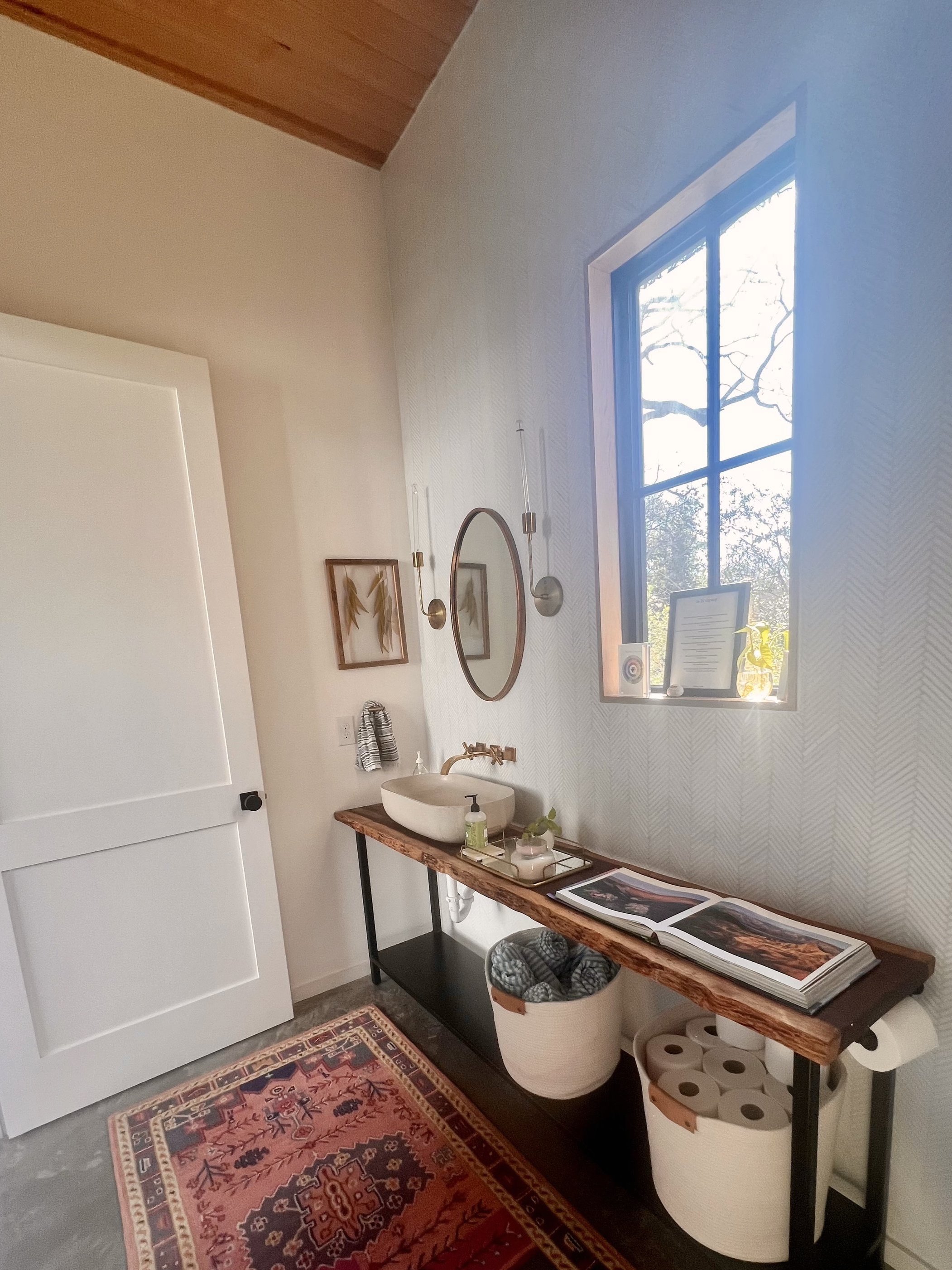 A bathroom with a wooden ceiling, a window with black framing, a round mirror, wall-mounted lights, a white basin sink, a rustic wooden counter with a soap dispenser, magazines, and decorative items, a woven basket with rolled towels, a laundry baske