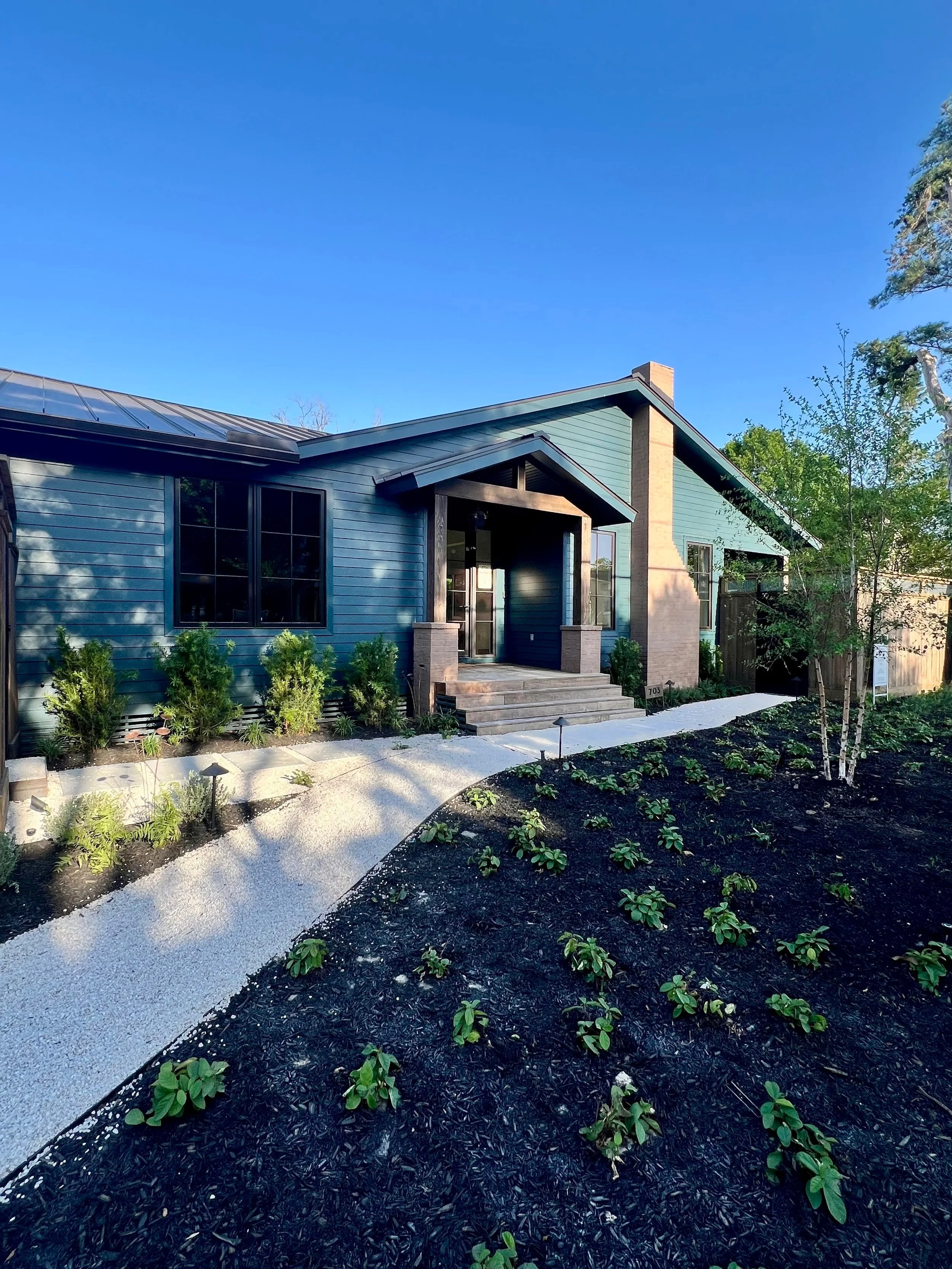 Newly built house with blue siding, black trim, a chimney, a covered front porch, a gravel walkway, burial planting beds, and a garden in the front yard under a clear blue sky.