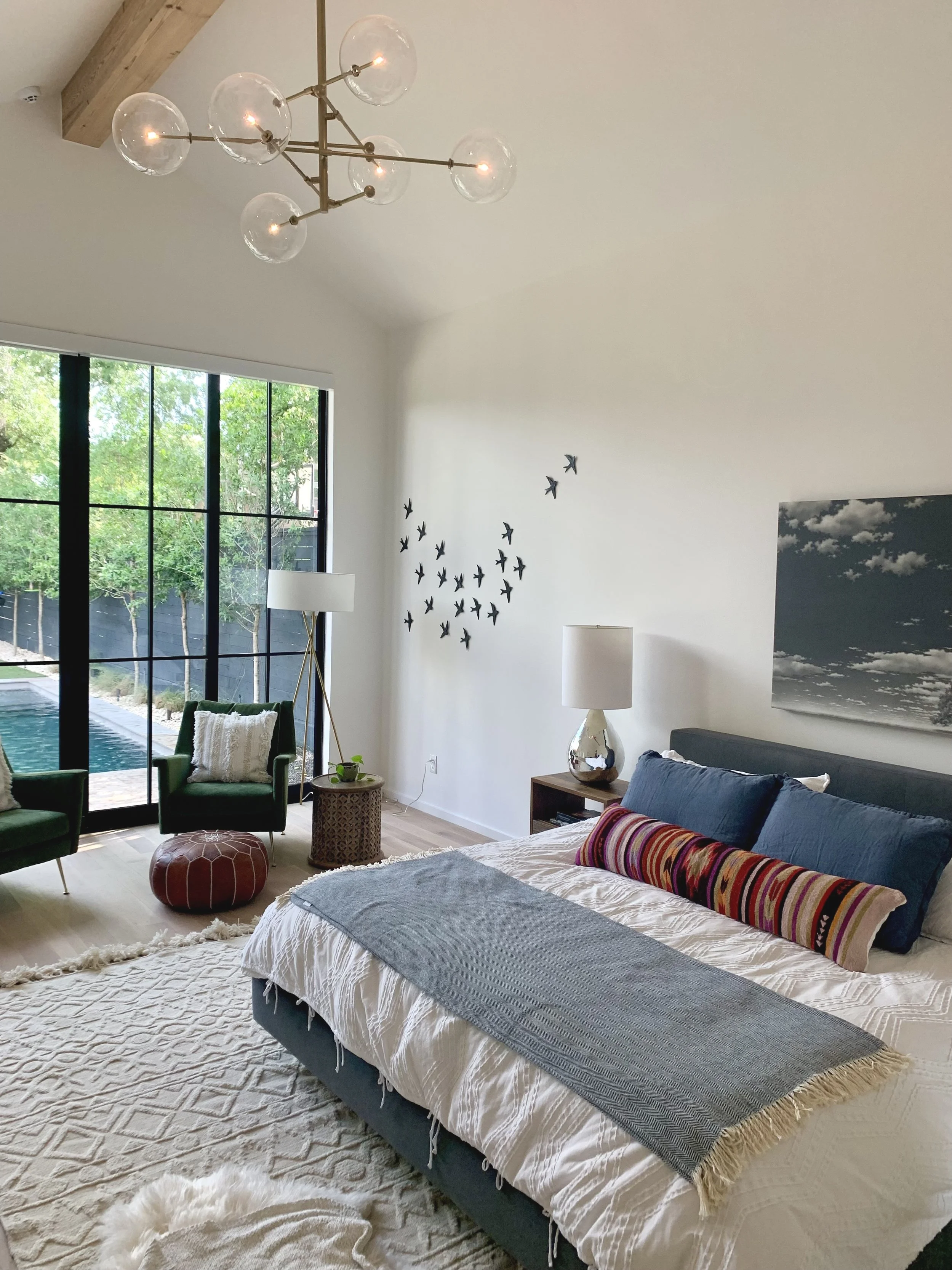 A bedroom with a large window showing a backyard pool and trees. The bed has white bedding with dark blue pillows and a long colorful striped pillow. There are two lamps on bedside tables, one with a white lampshade and the other with a ceramic base.