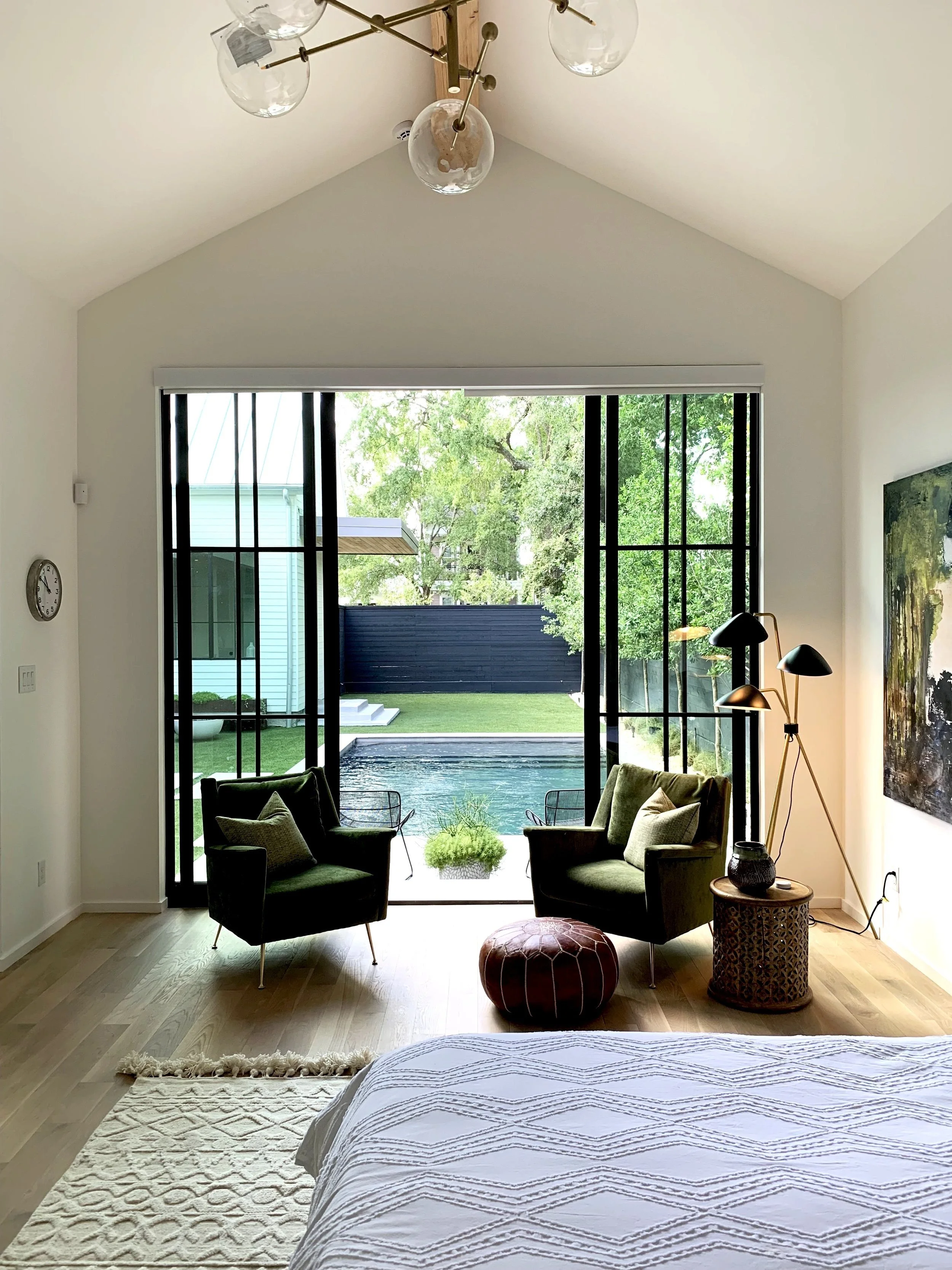 Modern bedroom with large sliding glass doors opening to a backyard pool and greenery, two green armchairs, a leather ottoman, a side table with a lamp, and a large painting on the wall.