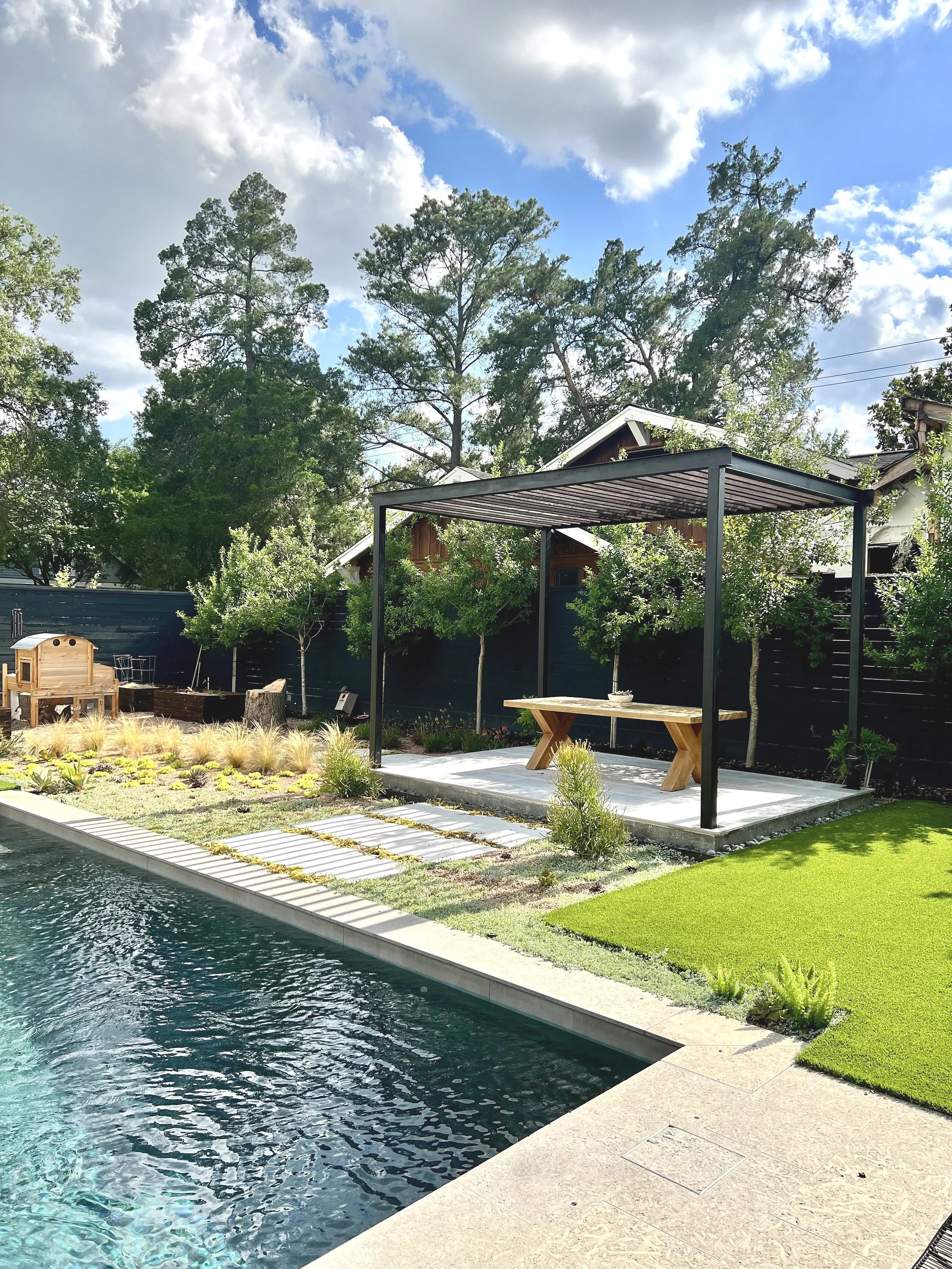 Backyard with swimming pool, covered patio with wooden table, trees, and a black fence under a partly cloudy sky.