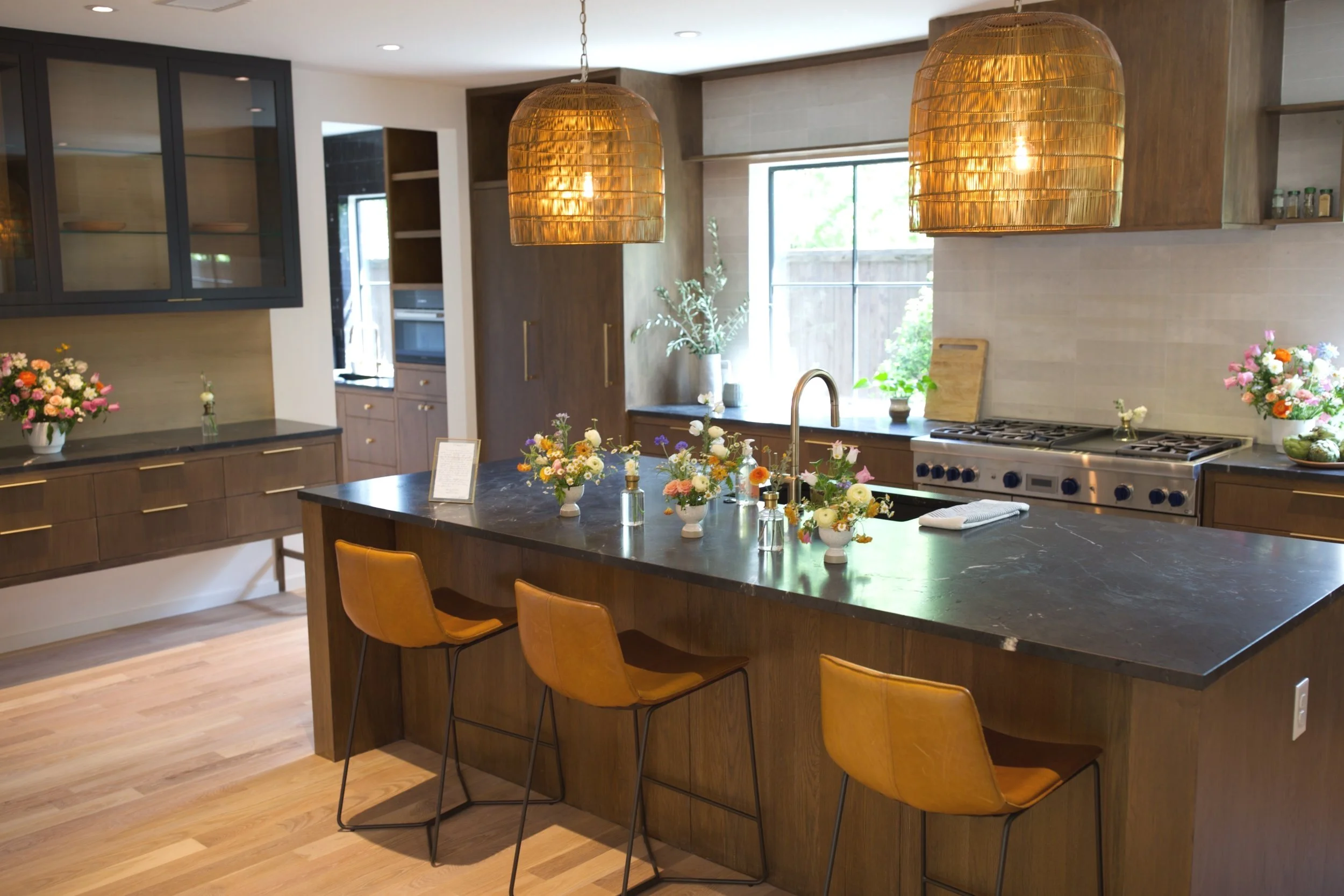 Modern kitchen with a dark marble island topped with flower arrangements, three brown chairs, wooden cabinets, a stove, and large woven pendant lights.