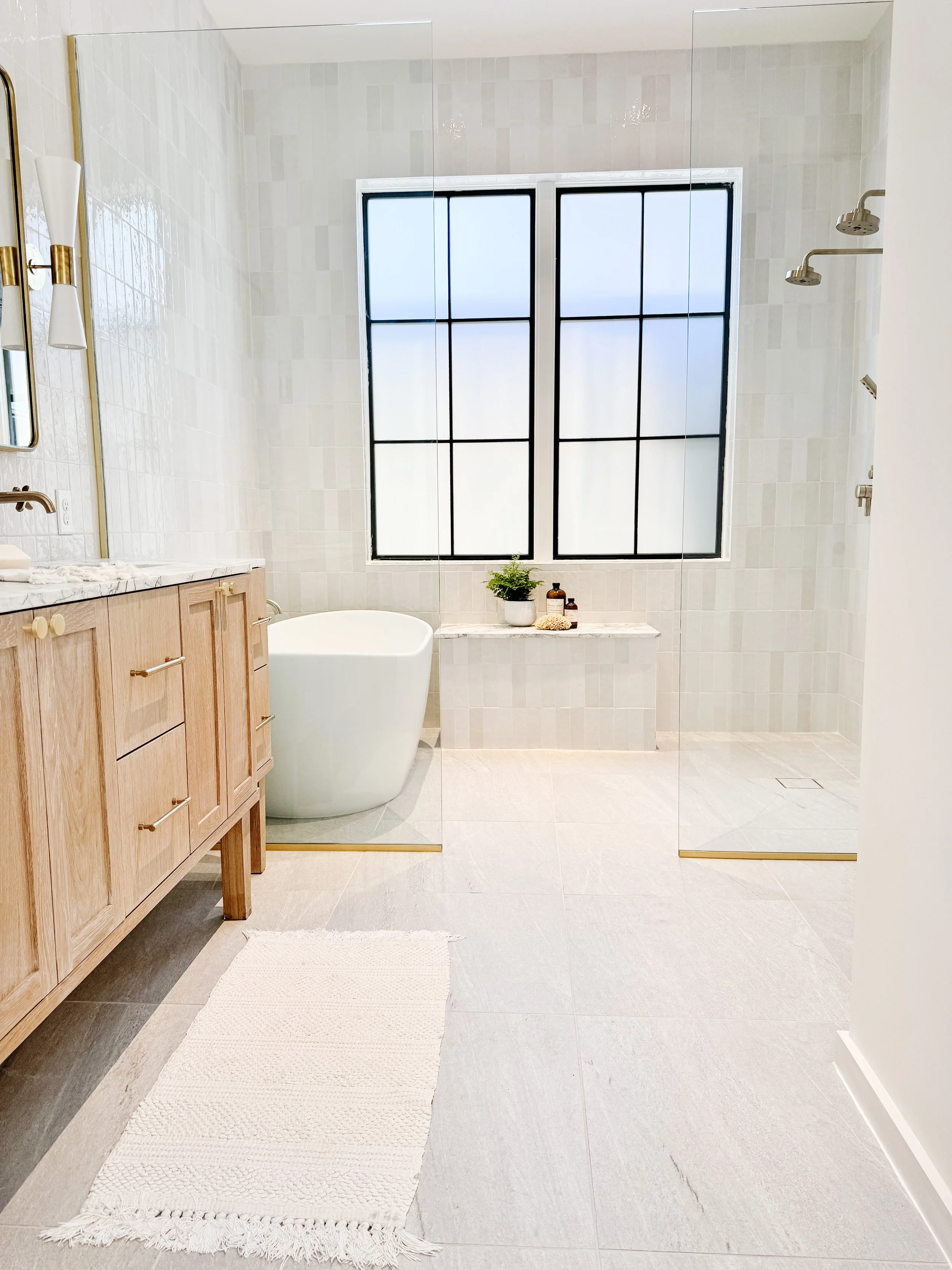 A spacious modern bathroom with a freestanding bathtub, large window with black grid frames, a wooden vanity with a marble countertop, and a walk-in shower with glass doors.