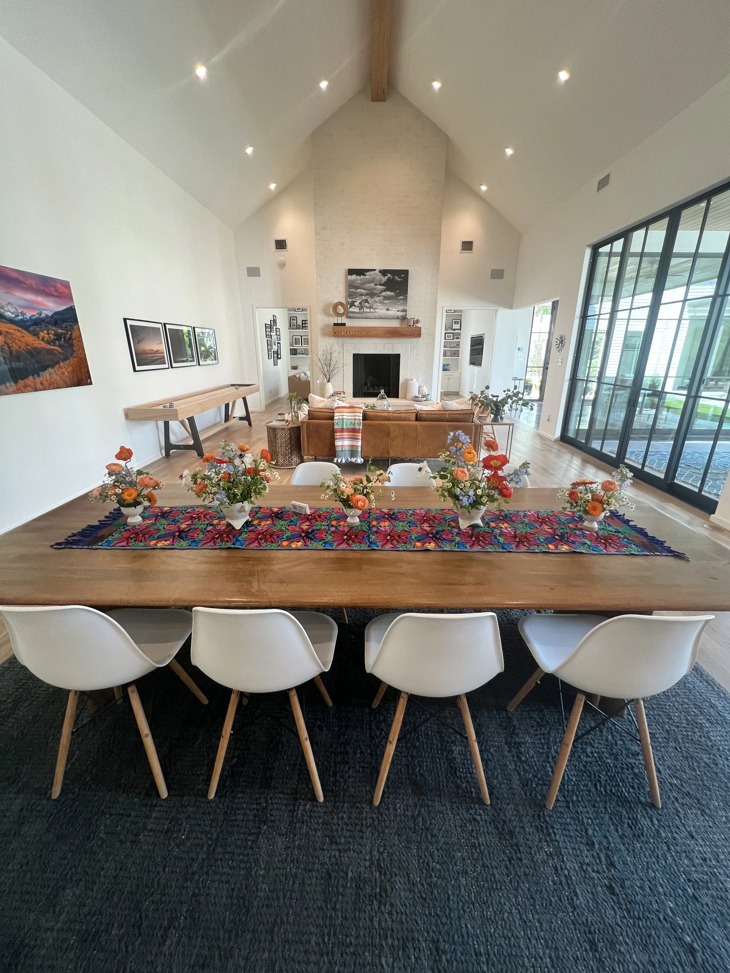 A spacious living room with a high vaulted ceiling, fireplace, and large glass doors. A wooden dining table with six white chairs is decorated with colorful flower arrangements. The room has light-colored walls and hardwood floors.