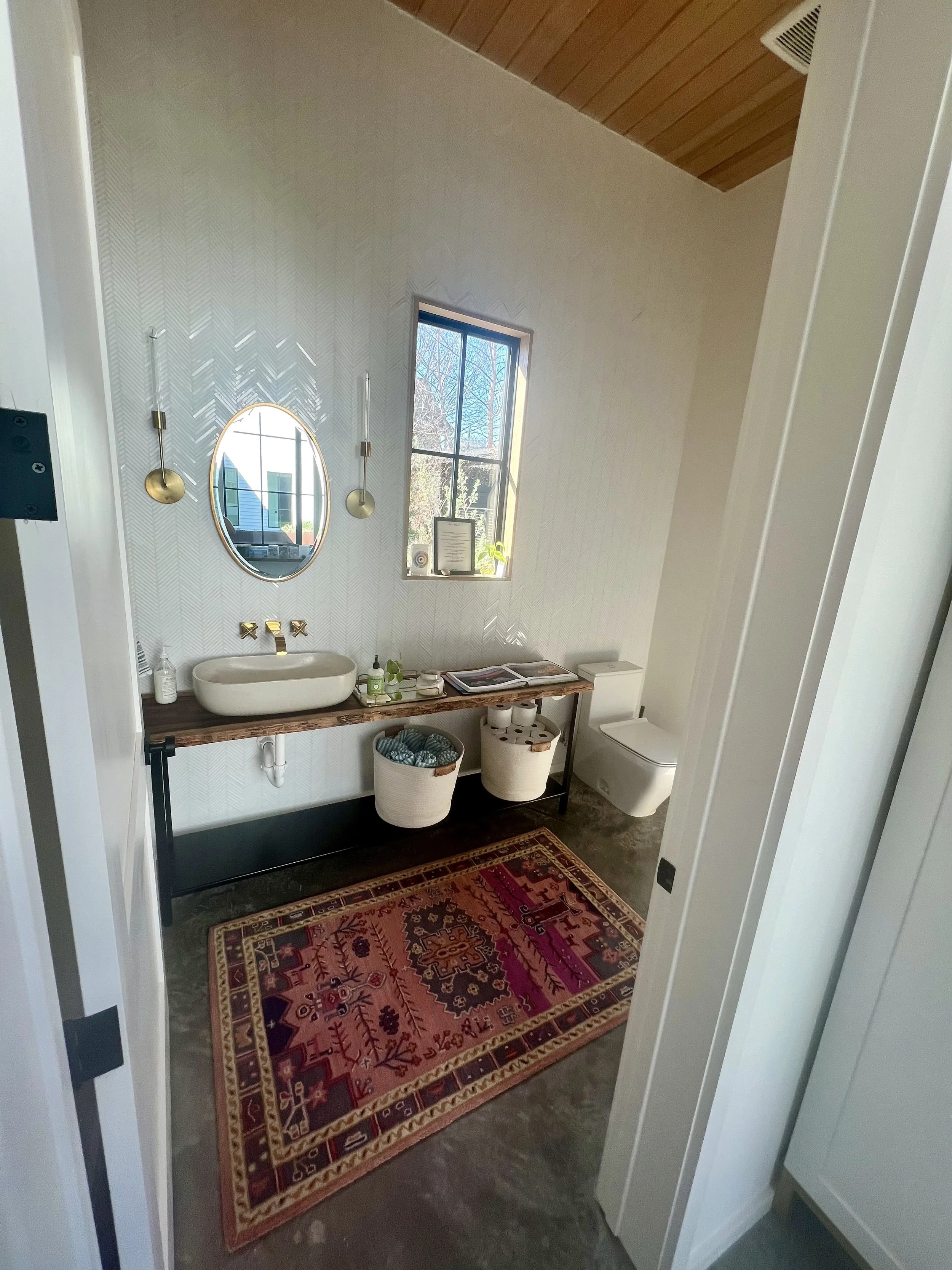 Modern bathroom with a rustic wooden vanity, a vessel sink, a round mirror, and hanging brass light fixtures. There is a window allowing natural light, with small potted plants and decorative items on the windowsill. A colorful Persian-style rug is o