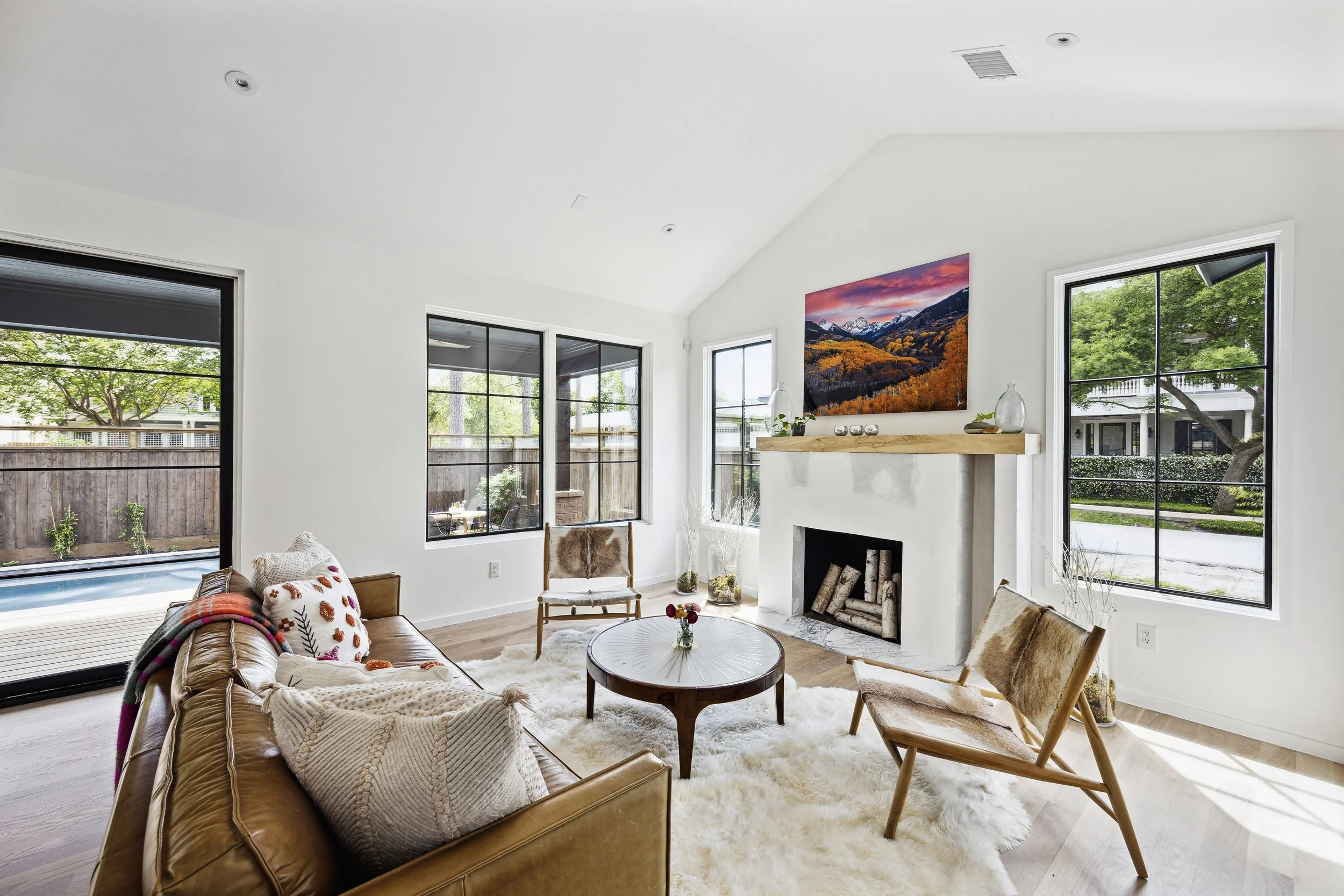 Bright living room with a leather sofa, a round coffee table, two wooden chairs with fur cushions, a fireplace, large windows, and a mounted flat-screen TV displaying a mountain landscape.