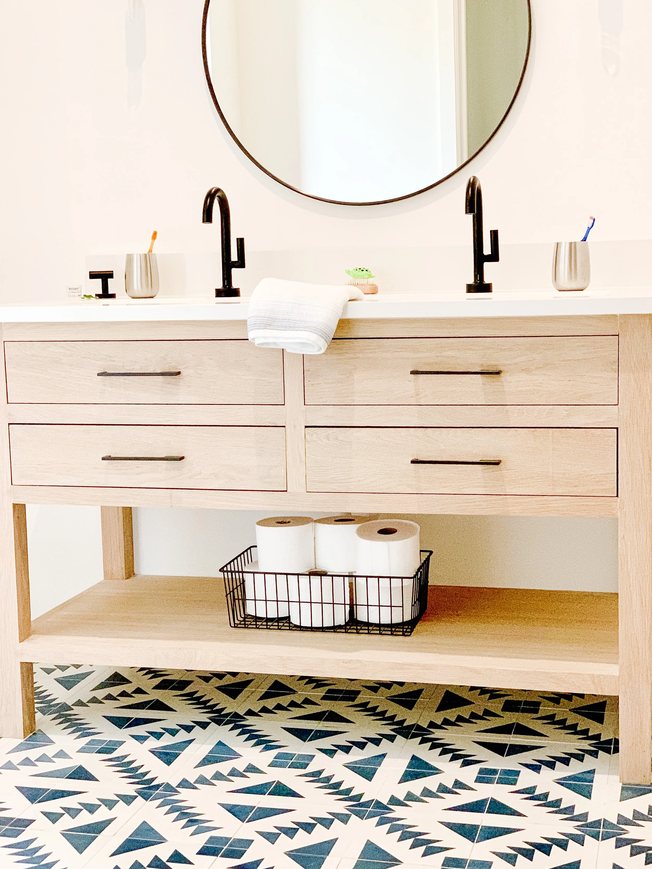 Bathroom vanity with two black faucets, a large round mirror, a white countertop, and light wood drawers. There are toothbrushes and cups on the counter, a white towel hanging over the countertop, and toilet paper rolls in a black wire basket below. 