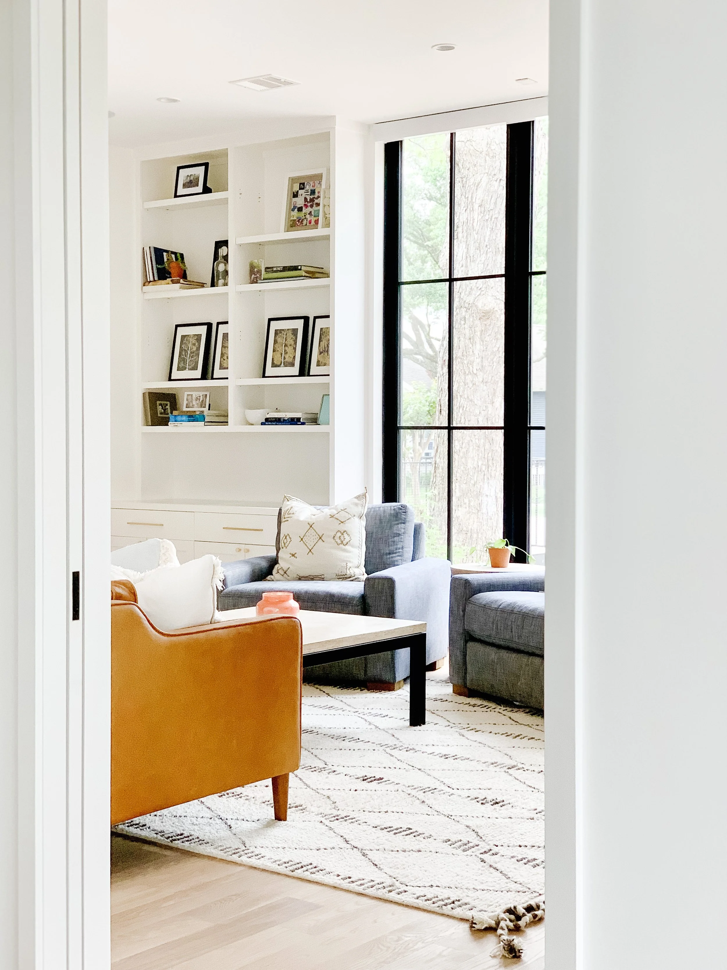 Bright living room with large window, dark gray armchair and sectional, orange armchair, coffee table, and white rug with geometric design.