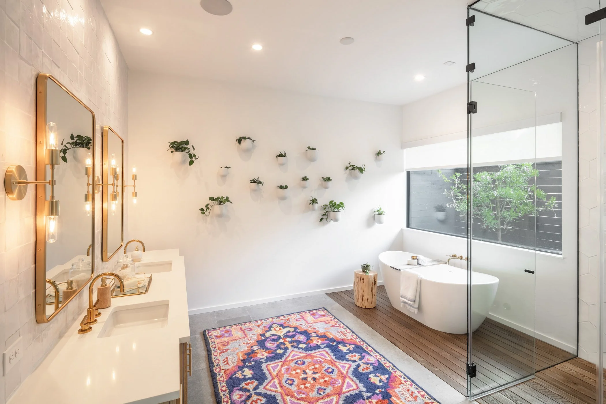Modern bathroom with white walls, wooden accents, and a large window. Features include two sinks with gold fixtures, decorative mirrors, wall-mounted plants, a colorful rug, a standalone bathtub, and a glass-enclosed shower area.