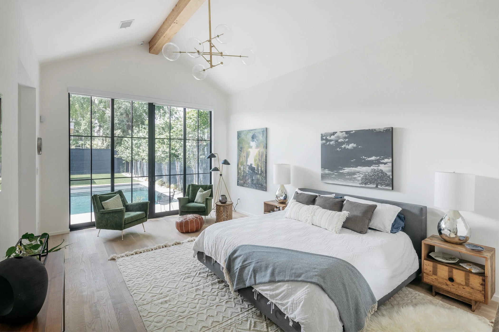 Modern bedroom with large window, white walls, wooden floors, bed with gray and white bedding, artwork, green chairs near window, and decorative lamps.