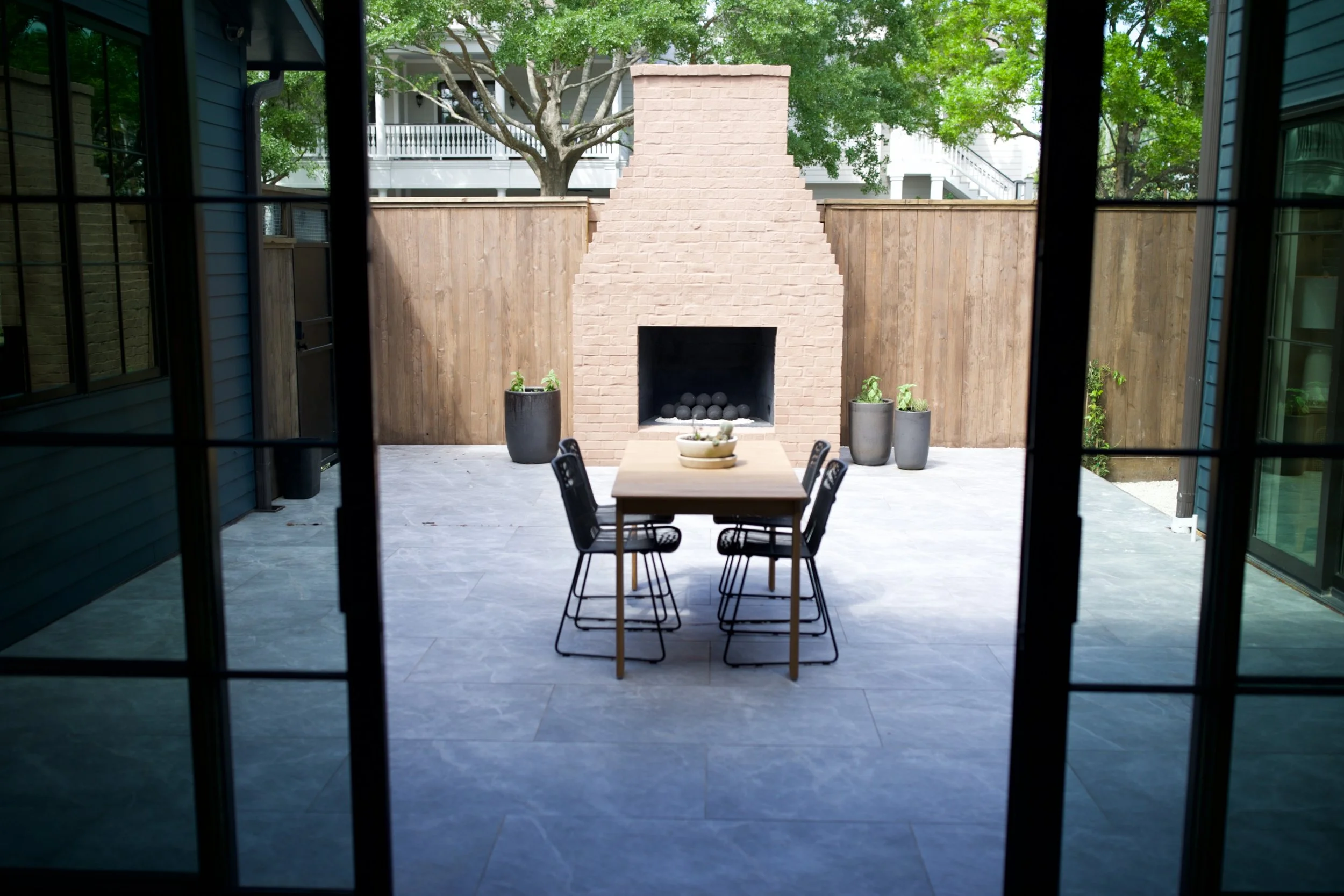 View of an outdoor patio with a brick fireplace, a wooden dining table, six black chairs, potted plants, and surrounded by a wooden fence.
