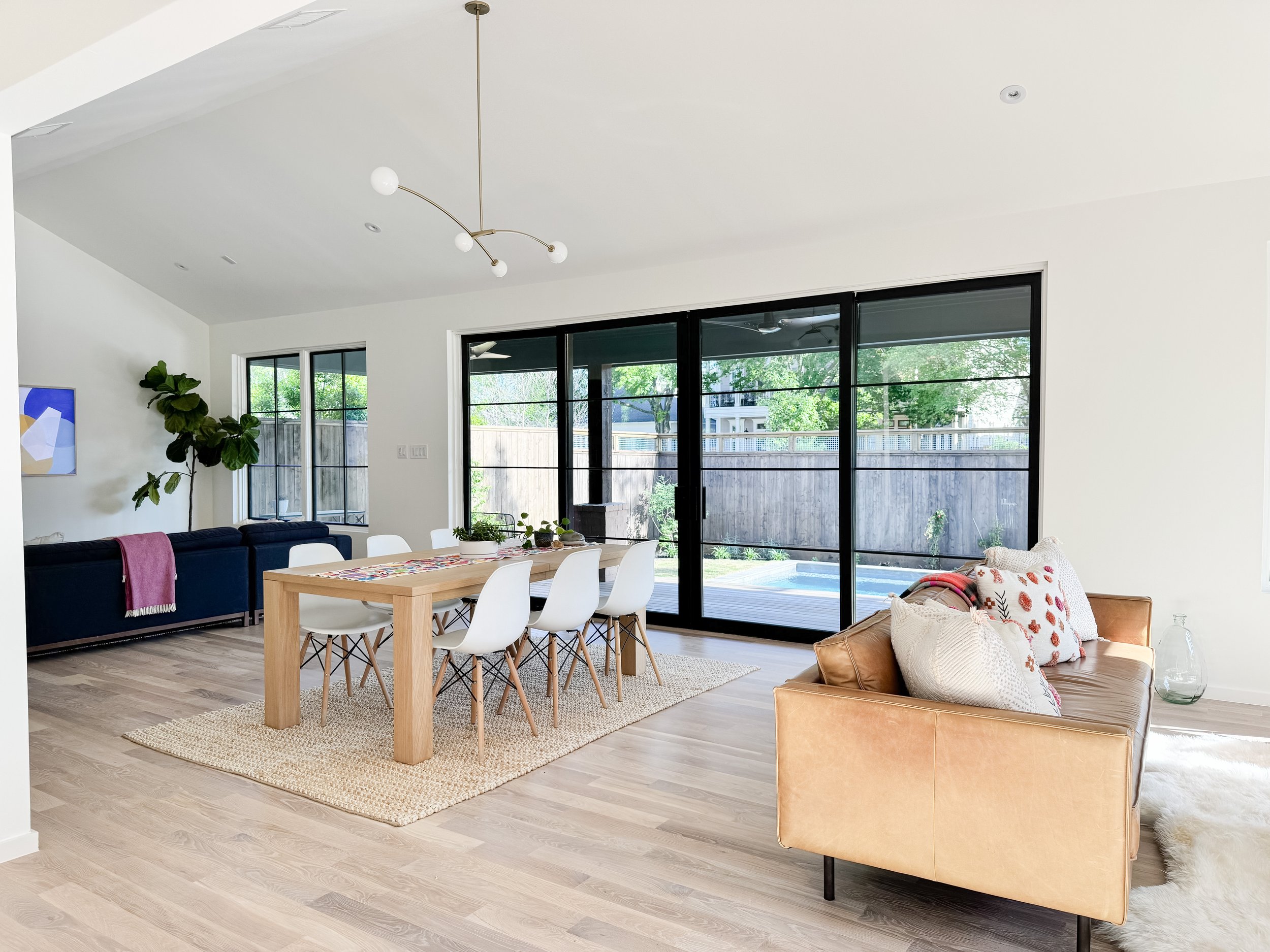 Bright living and dining room with large sliding glass doors, a light wood dining table with six white chairs, a tan leather sofa with pillows, a modern chandelier, and a view of a backyard with a fence and trees.