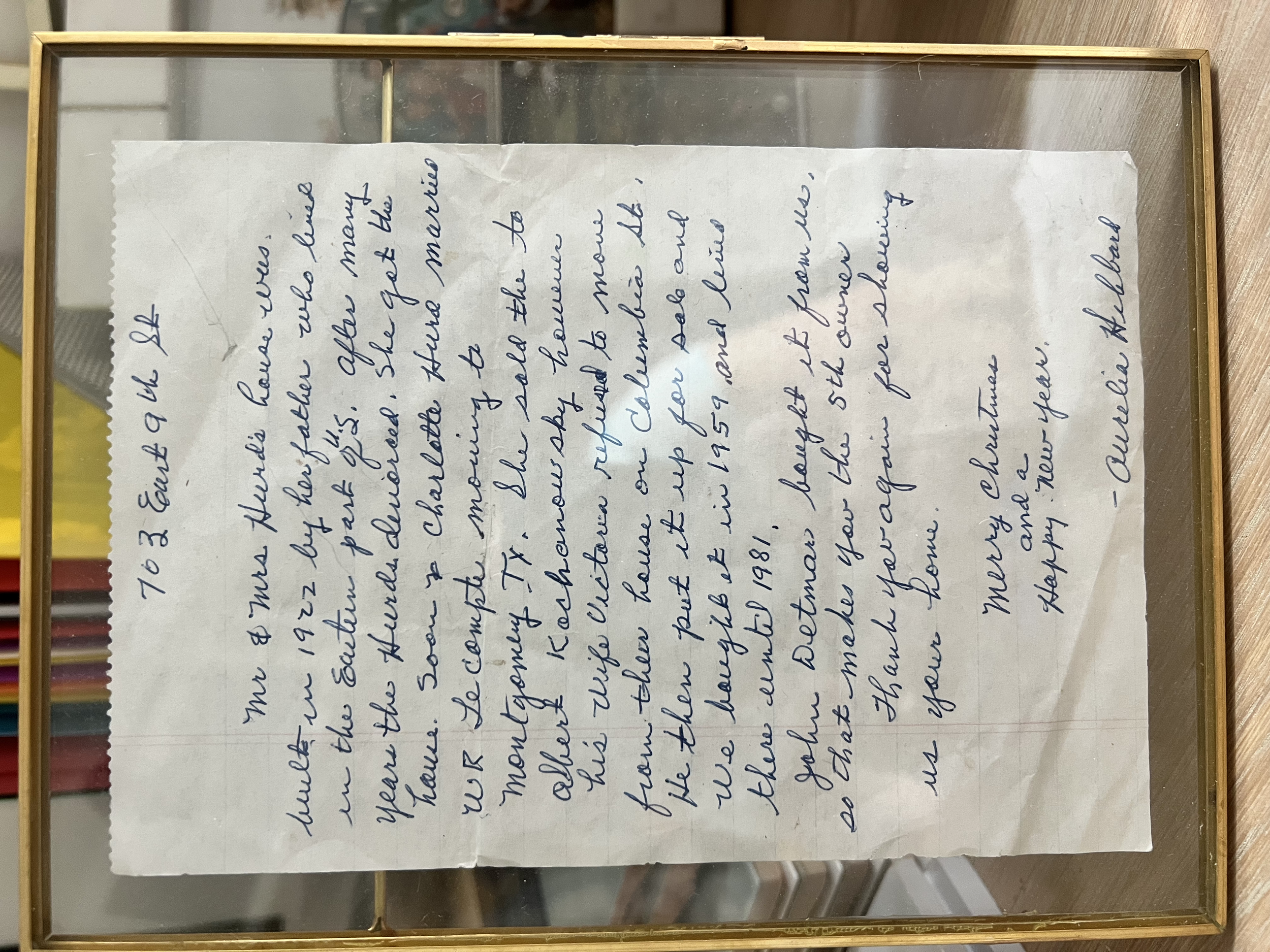 A handwritten note in cursive on a white sheet of paper, framed behind glass with a gold border, resting on a wooden surface.