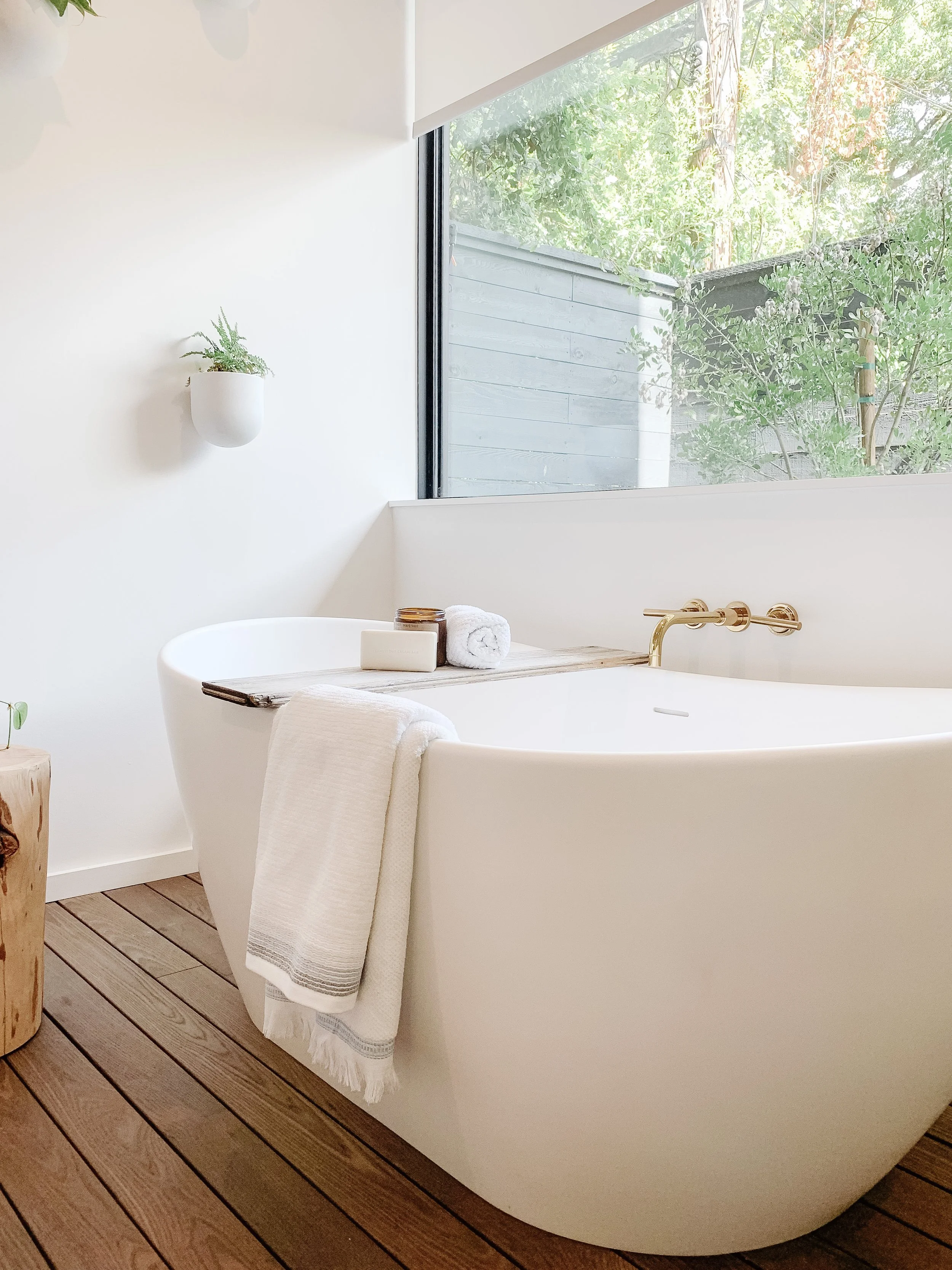 Modern white bathtub with gold fixtures in minimalist bathroom with wooden floor and large window showing green outdoors.