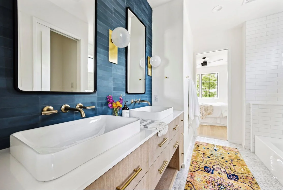 Modern bathroom with a dual vanity, blue accent wall, white vessel sinks, large mirrors, gold fixtures, wall sconces, a colorful rug, and a view into a bright bedroom.