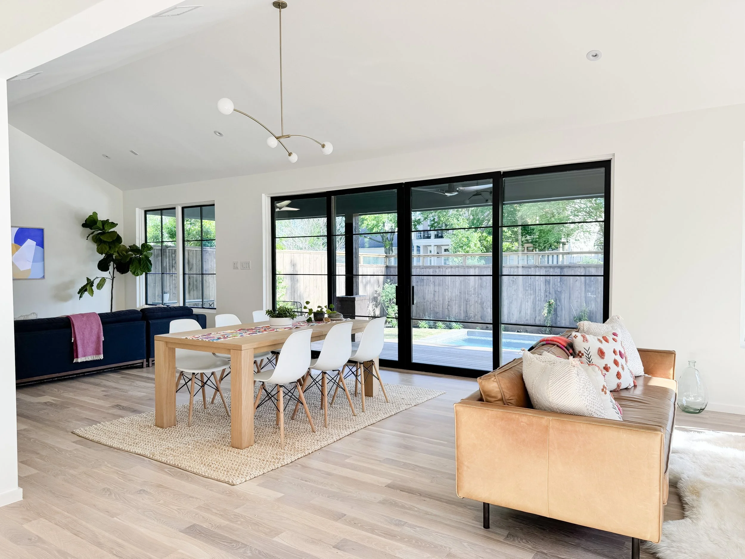 Bright open-concept living and dining area with light hardwood floors, white walls, large sliding glass doors leading to a backyard with a pool, a wooden dining table with white chairs, a blue sofa with a pink throw blanket, and a tan armchair with t
