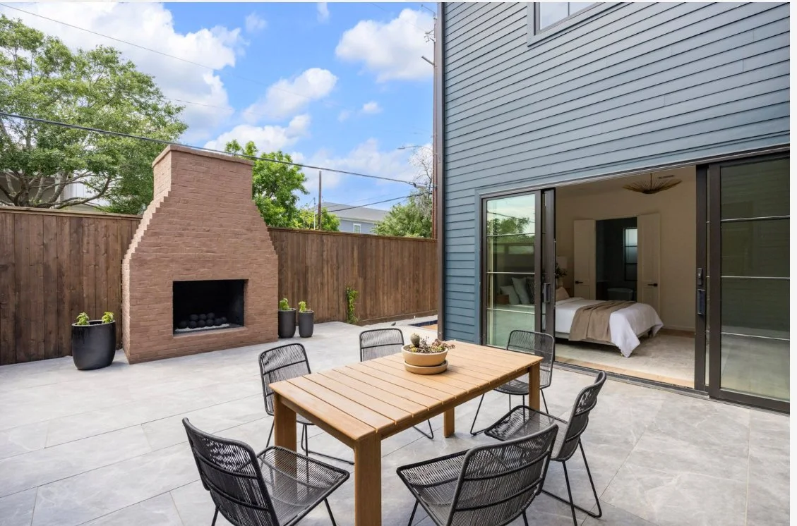 Outdoor patio with a brick fireplace, a wooden dining table with six black chairs, and potted plants. Open sliding glass doors leading to a bedroom inside the house.