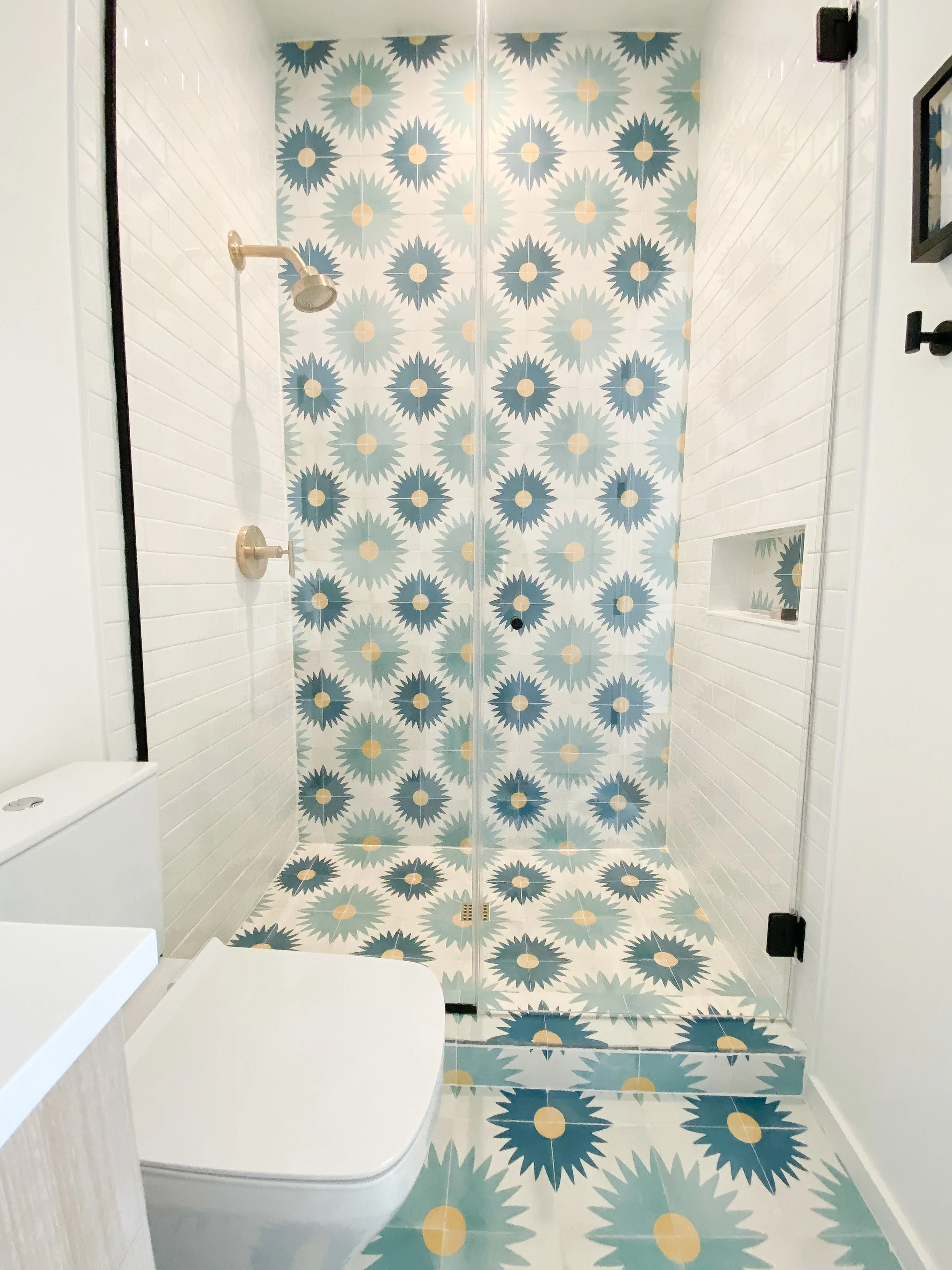 Modern bathroom with a shower featuring blue and yellow patterned tiles on the walls, floor, and door. Includes a white toilet and white walls with a small built-in shower niche.