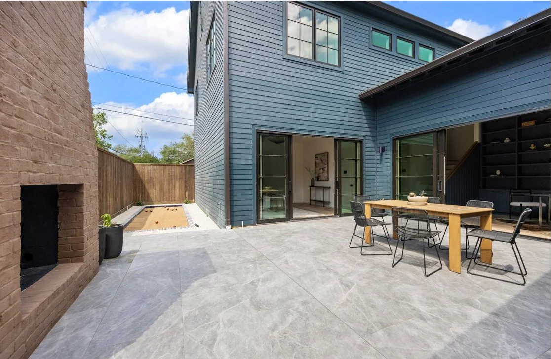 Backyard patio with gray stone flooring, a wooden dining table with four black chairs, a small sandbox area with a wooden fence, and a blue two-story house with large glass doors and windows.