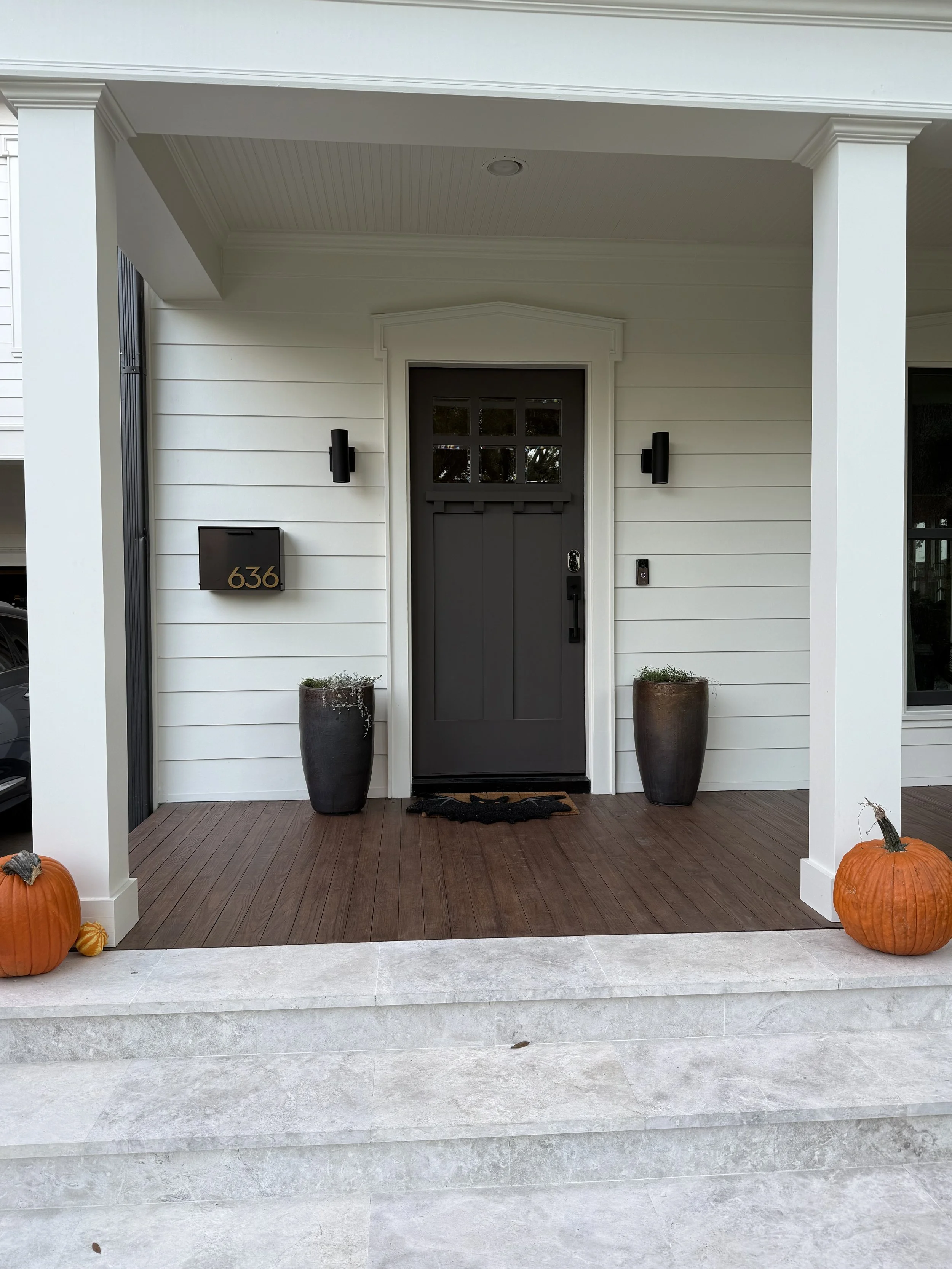 Front steps of a house decorated for fall with pumpkins, black doormat, potted plants, black mailbox with gold house number 636, black entrance door, and outdoor wall lights.