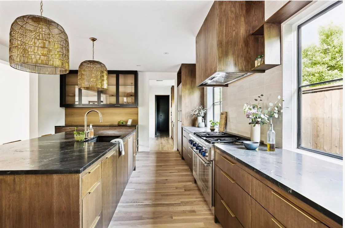 Modern kitchen with wooden cabinets, marble countertops, two hanging woven pendant lights, and flower vases near a large window