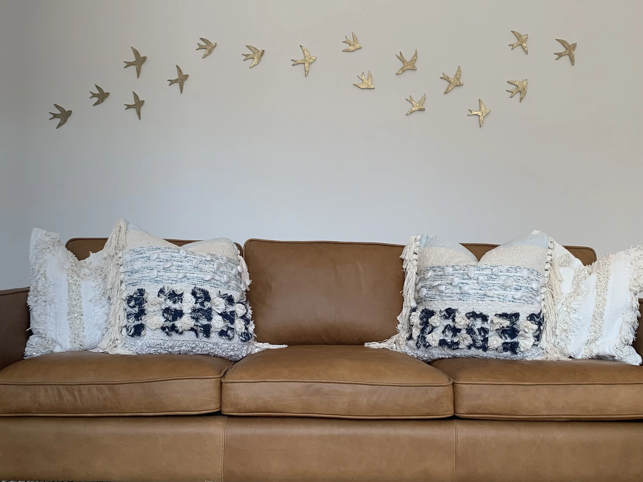 A brown leather sofa with three decorative pillows featuring navy and cream embroidery and fringe, set against a blank wall decorated with metallic bird wall art.