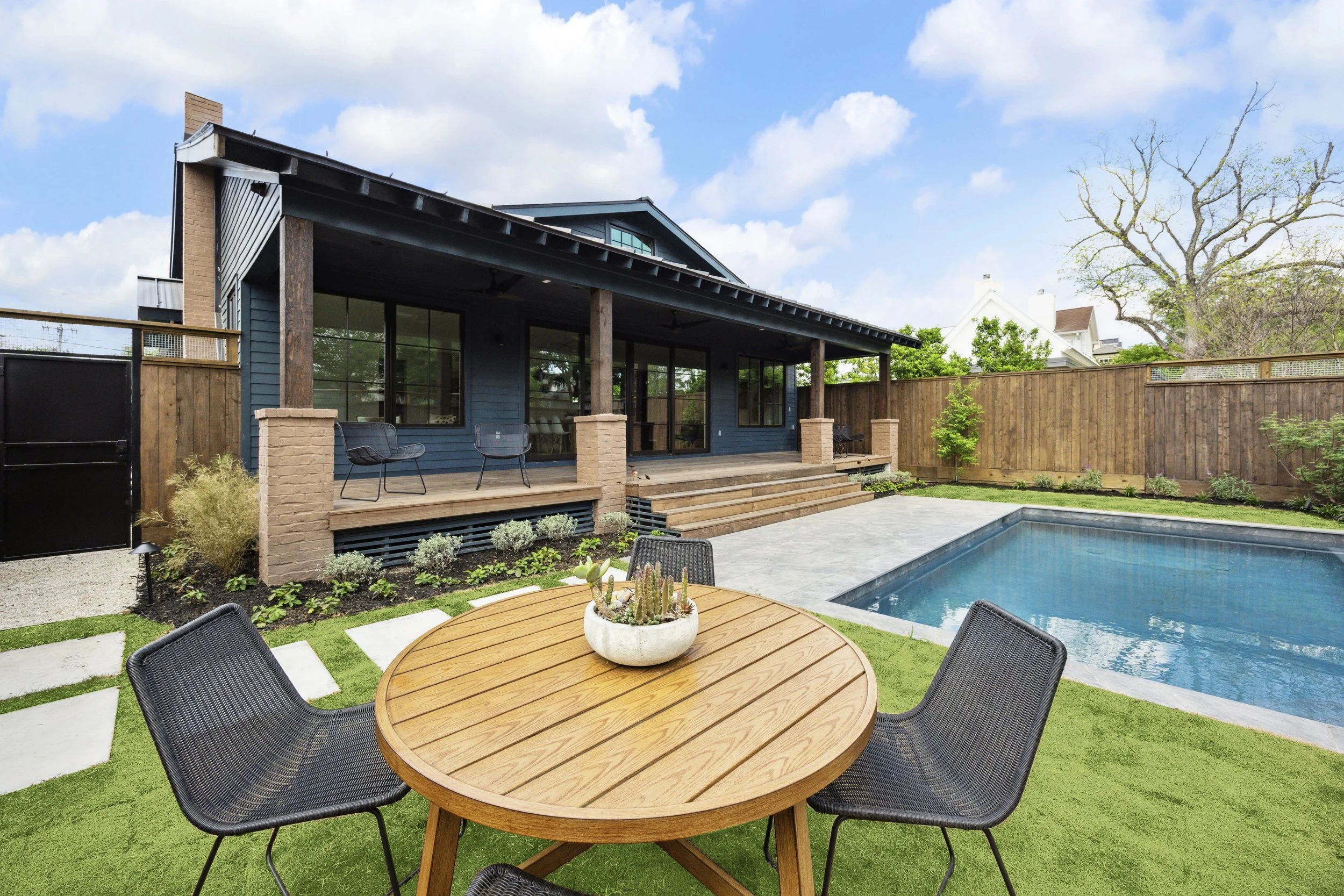 Backyard with a wooden deck, a swimming pool, a round wooden table with black chairs, and a modern house with large window panels, wooden steps, and a fenced yard under a partly cloudy sky.