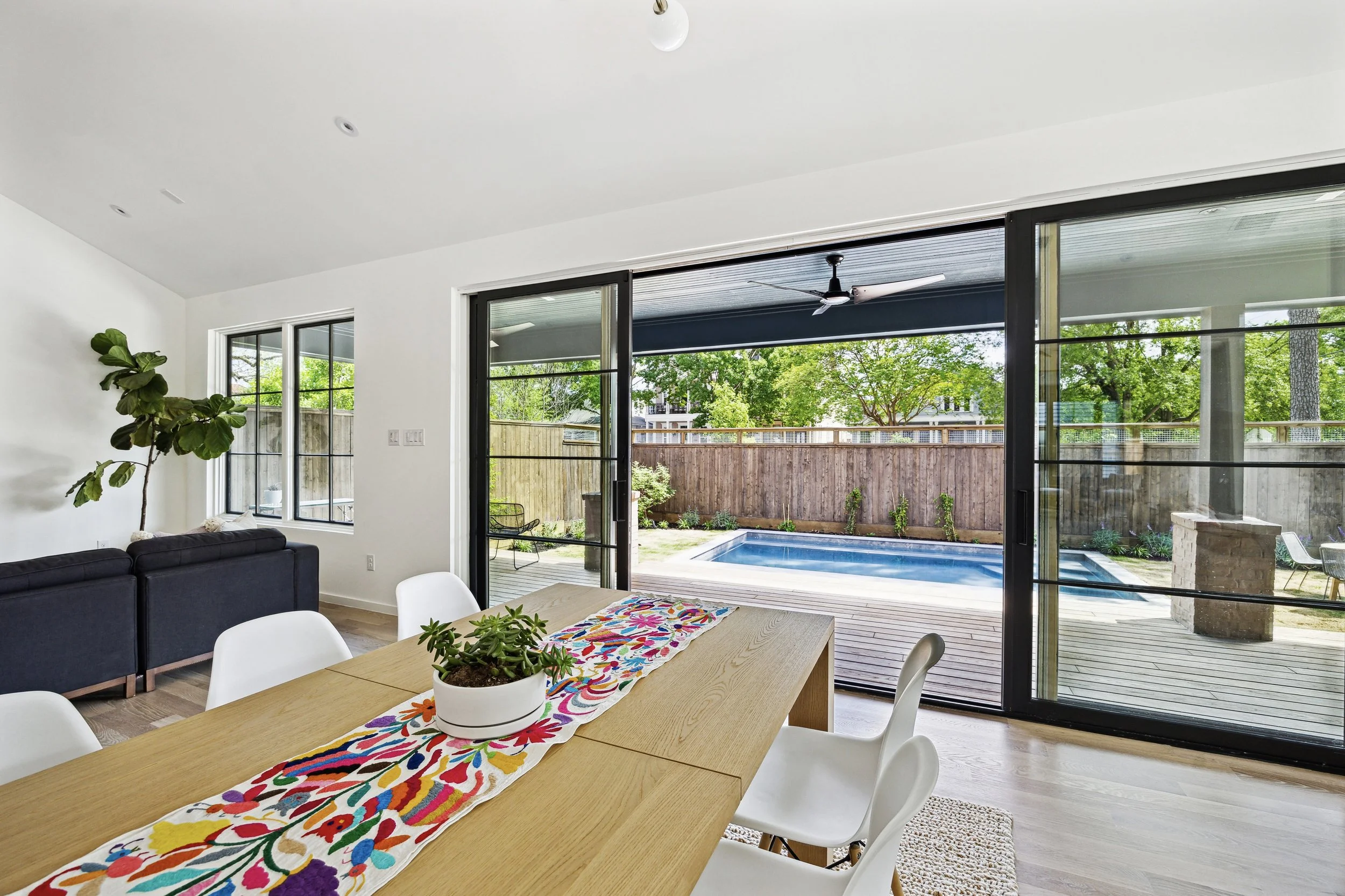 Dining room with a wooden table, white chairs, and a potted plant centerpiece, opening to a backyard patio with a pool, wooden deck, and green trees.