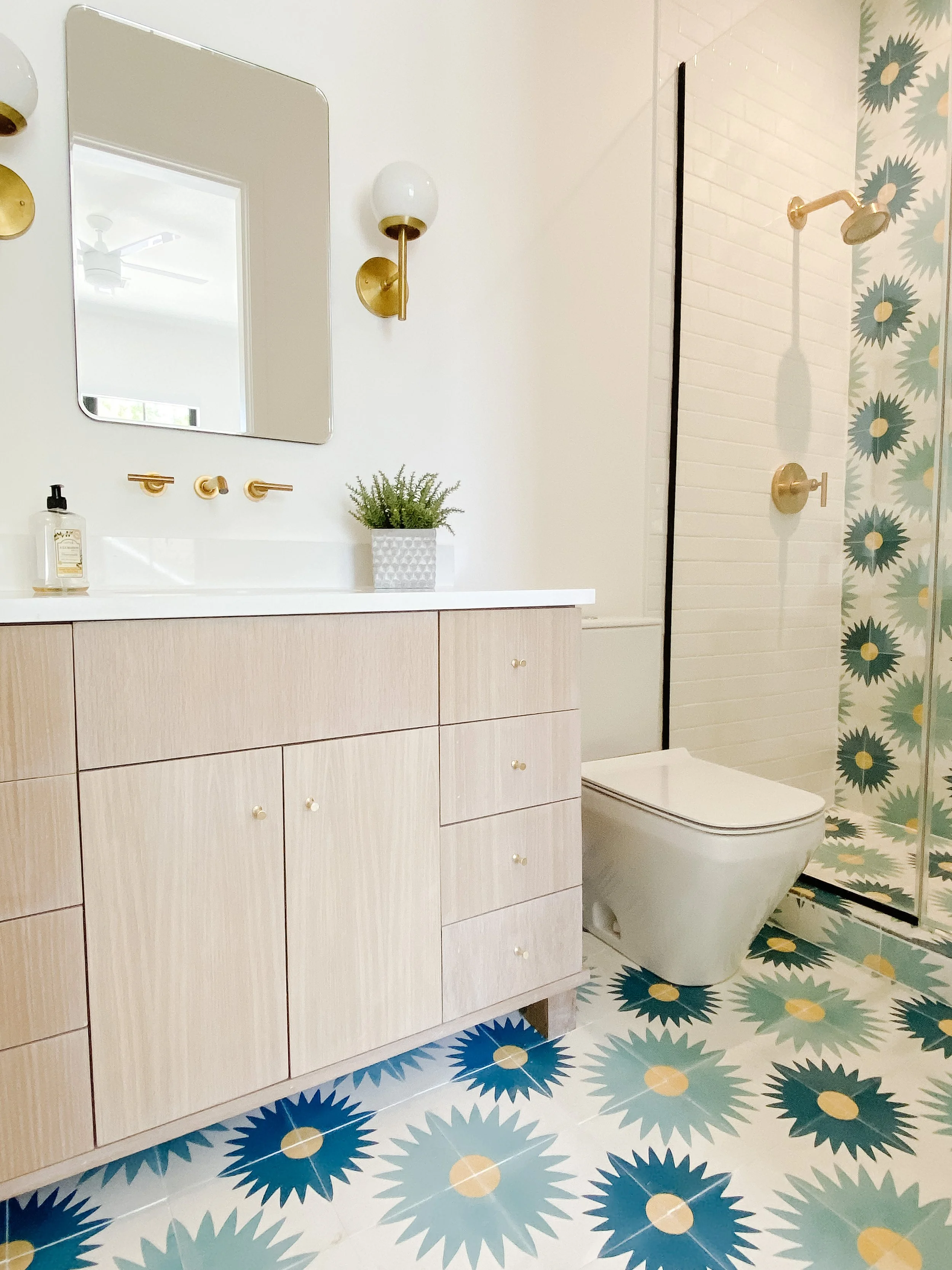 Modern bathroom with a white vanity, gold fixtures, a square mirror, a wall-mounted plant, a toilet, and a shower with a floral pattern wall and floor tiles in blue, green, and beige colors.
